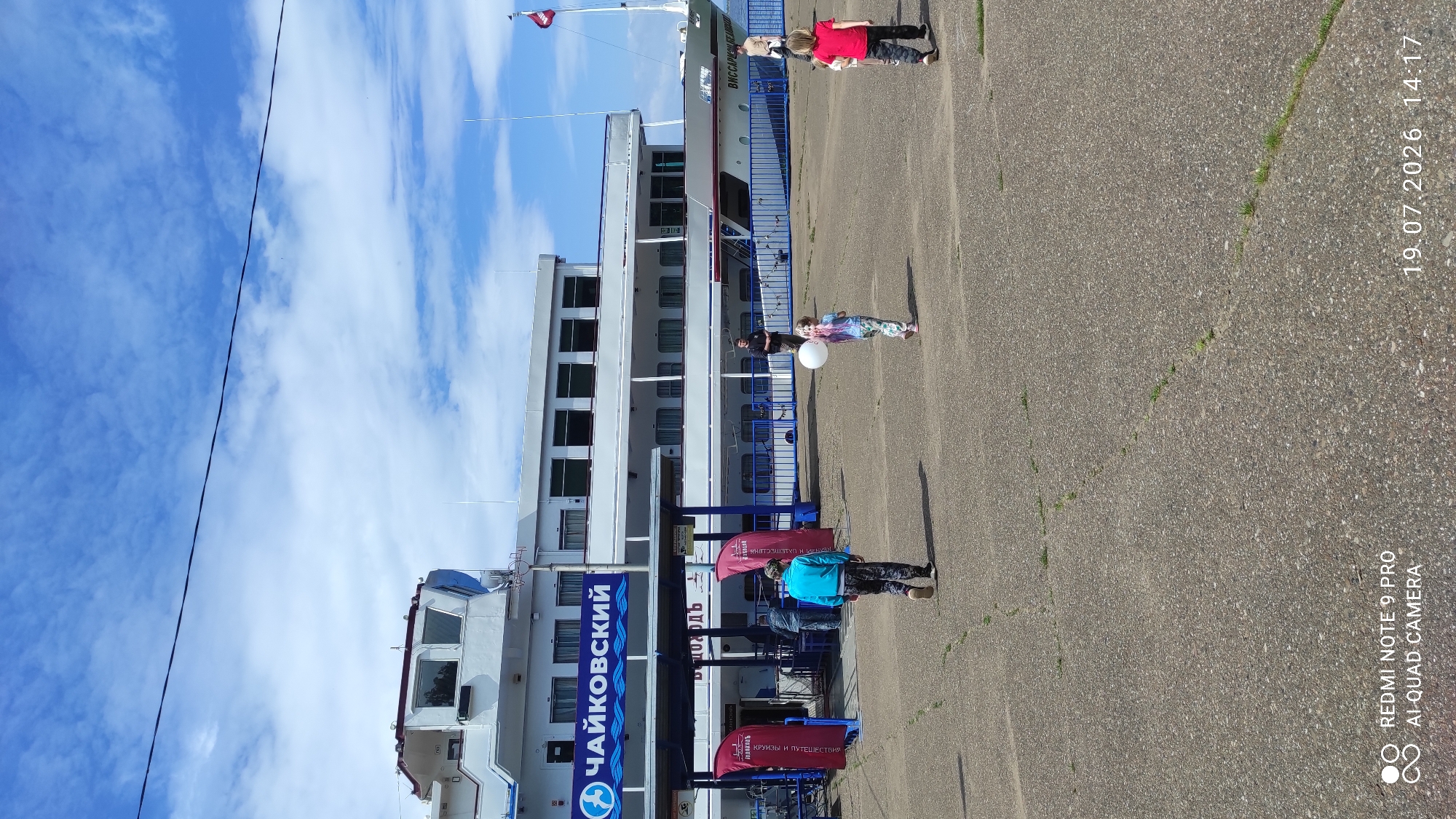 The image depicts a bright, sunny day at the riverfront in Chaikovsky, Russia, specifically at the Chaikovsky River Port. In the background, a large white passenger ship, identified by the name "Чайковский" (Chaikovsky) on its side, is docked. The ship has multiple decks and windows. In front of the ship, there's a covered walkway with blue railings and red banners. Several people are visible on the paved area in the foreground. On the left, a person wearing a blue jacket and dark patterned pants walks away from the camera. To the right of the walkway, a person in a black shirt and pants stands. Further to the right, two children are walking. One child, wearing a colorful outfit, holds a white balloon. Another child, wearing a red t-shirt and dark pants, is also walking away from the camera. The ground is a grey, textured asphalt surface with visible cracks and some sparse grass. The sky is blue with scattered white clouds. The timestamp "19.07.2026 14:17" is visible in the bottom right corner, along with the camera information "REDMI NOTE 9 PRO AI-QUAD CAMERA."
