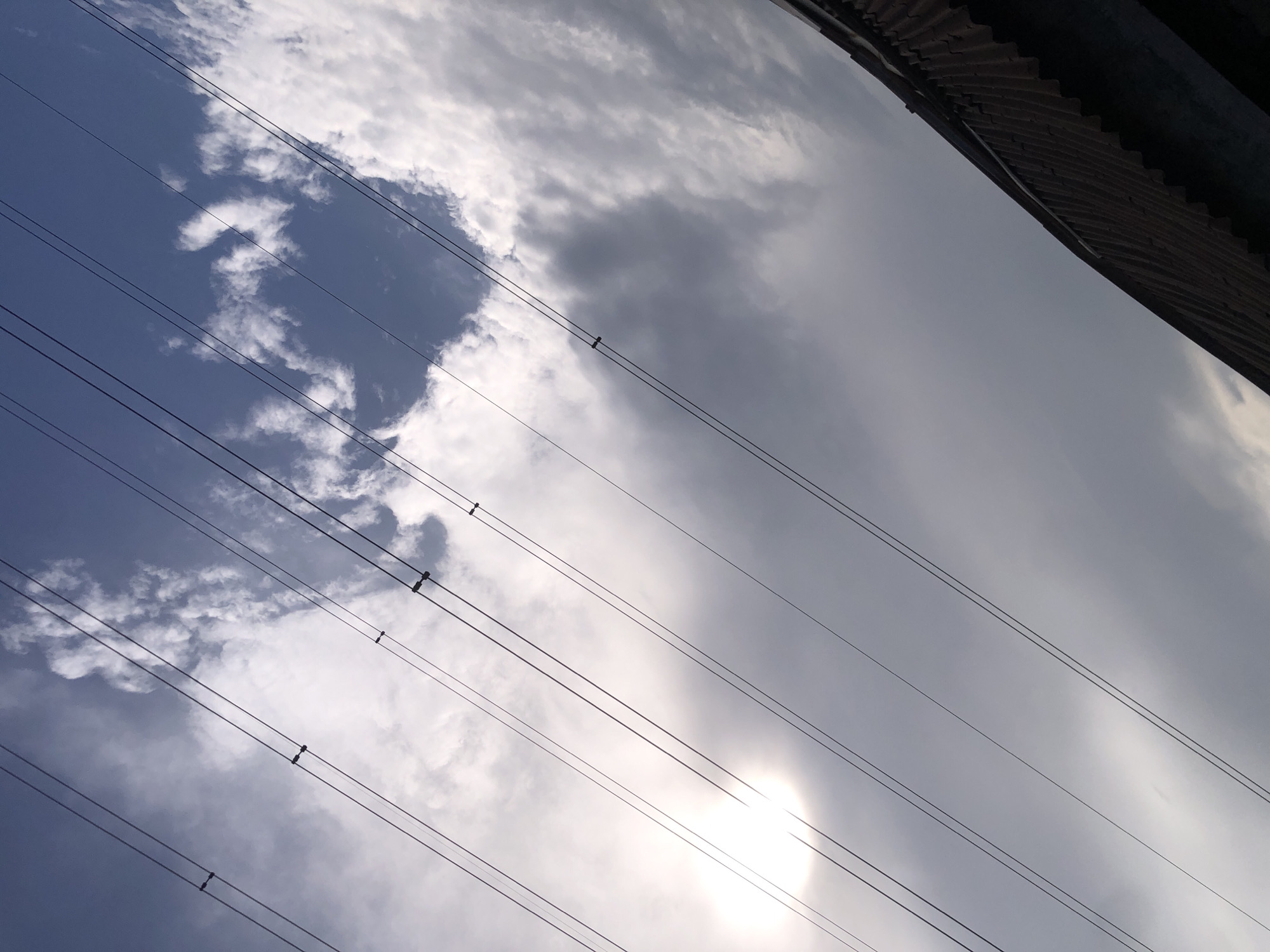The image depicts a close-up, low-angle shot looking upwards towards a cloudy sky. Several parallel wires, likely power lines, stretch diagonally across the frame from the lower left to the upper right. The sun is visible as a bright, diffused light source in the upper left quadrant, casting a halo effect through the clouds. The clouds are a mix of white and grey, suggesting varying densities. In the lower right corner, the corrugated metal roof of a building is visible, adding a textured, dark element to the composition. The scene appears to be captured outdoors, possibly in an urban or semi-urban environment given the presence of power lines and a building roof. The image does not contain any people or specific identifiable landmarks of Ado, Nigeria.