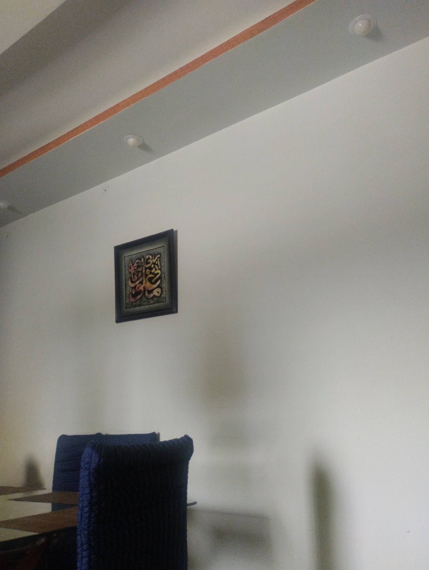 An indoor shot captures a section of a wall and ceiling. On the wall, a framed piece of artwork with calligraphy is displayed. Below the artwork, two dark blue chairs are partially visible, suggesting a seating area. The ceiling features a decorative element with an orange stripe and a few recessed light fixtures. The overall lighting is soft, with shadows cast on the wall. The location is not specified.