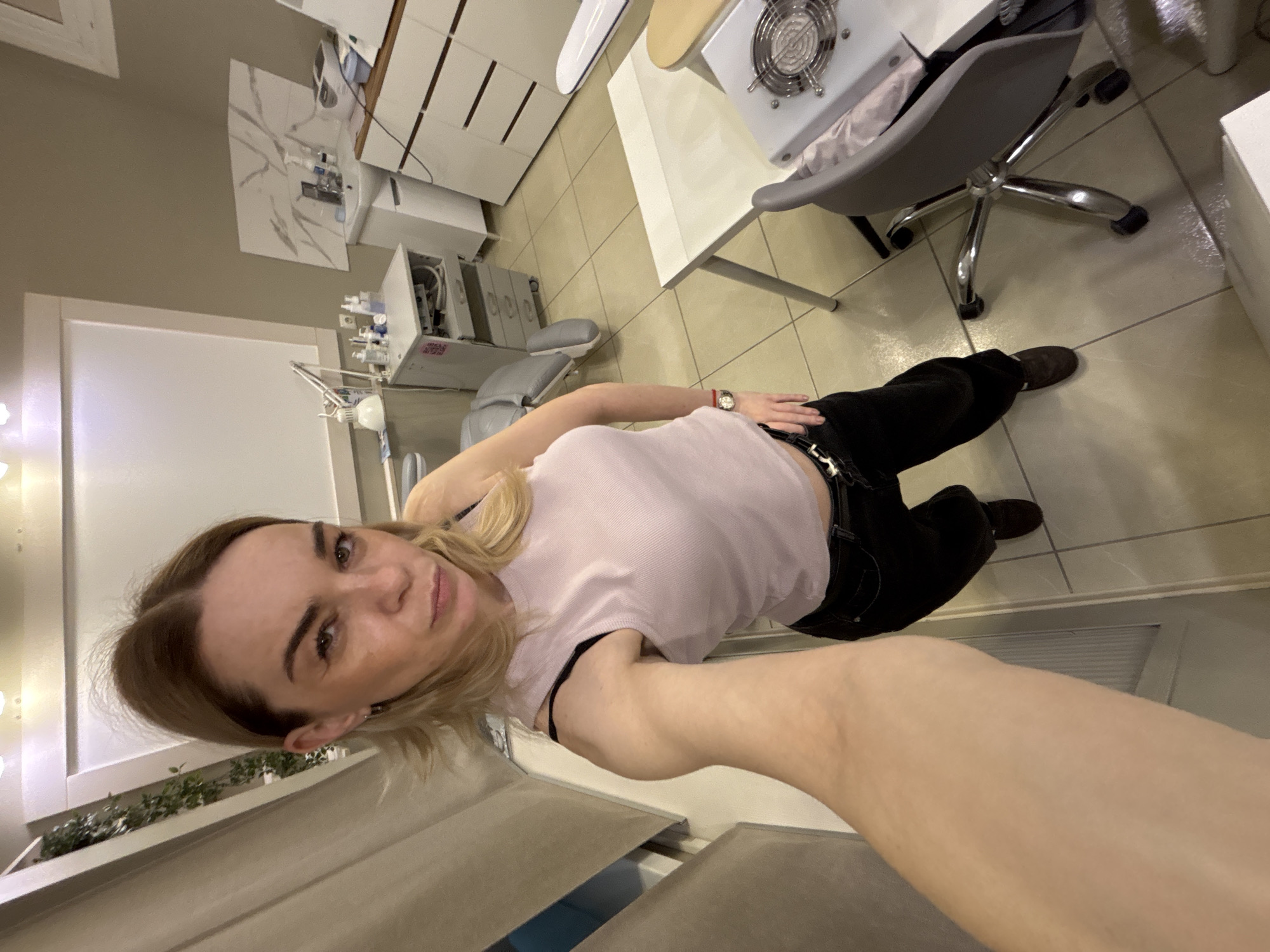 A selfie taken by a Caucasian woman in what appears to be a beauty salon or nail salon. She is wearing a light pink top and black pants. The background includes manicure stations, chairs, and various salon equipment. The location is undetermined.