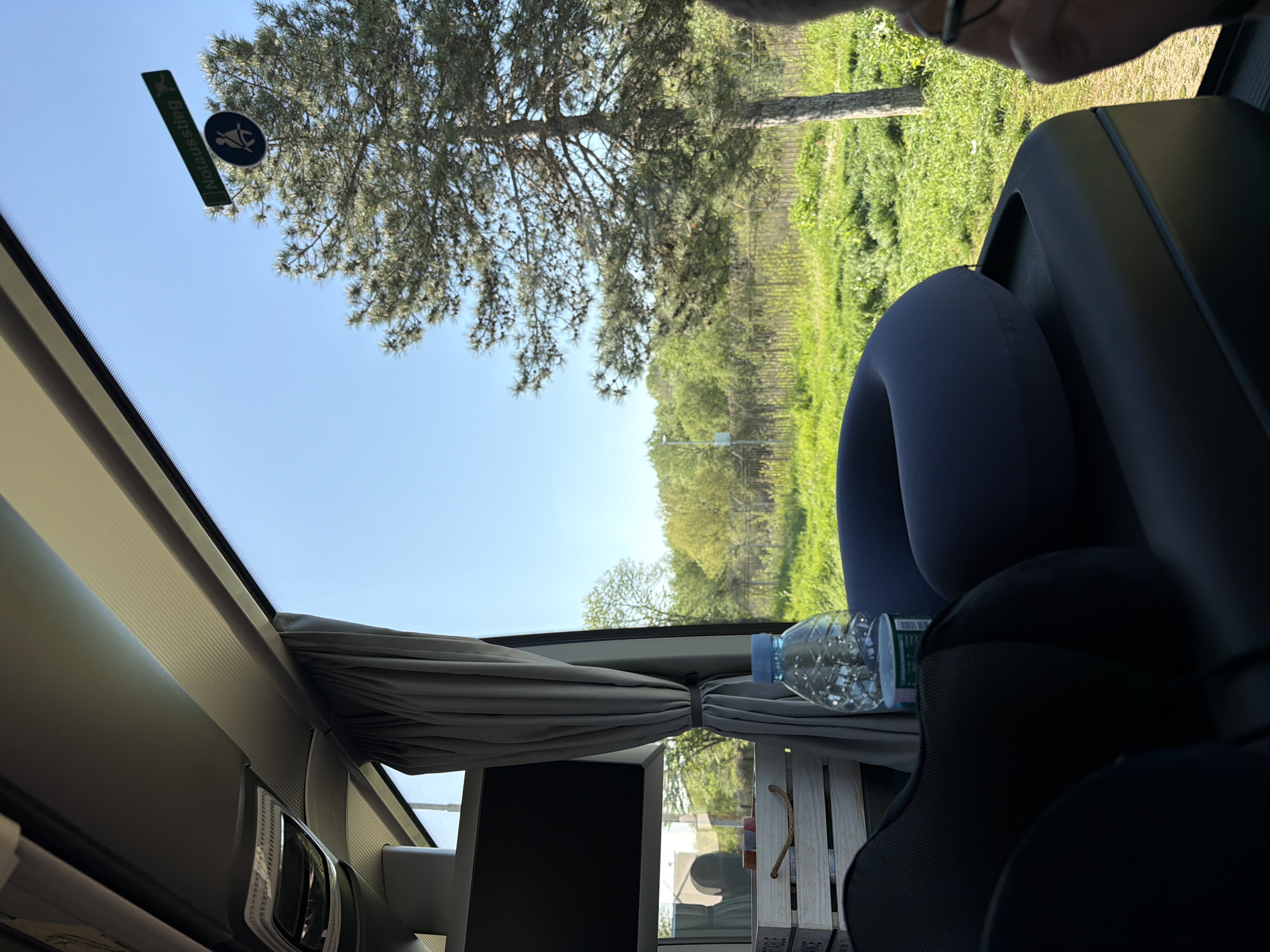 This image captures a bright, sunny outdoor scene viewed from inside a vehicle, likely a bus, campervan, or train. The perspective is from a passenger looking out a large window, with the image rotated 90 degrees clockwise from its natural orientation.
Through the window, a clear, light blue sky dominates the upper portion. A large pine tree with visible cones stands prominently against the sky. Beyond the pine tree, a landscape of lush green grass and dense foliage stretches into the distance under the bright daylight. Subtle linear patterns within the green area suggest a fence line or possibly rows of cultivation.
Inside the vehicle, a grey curtain is tied back to the side, framing the exterior view. To the right, a dark blue inflatable neck pillow rests on a black seat or headrest, hinting at travel comfort. A clear plastic water bottle with a blue cap sits on a surface beside the pillow, which also includes a portion of a wooden crate or tray. A black screen or monitor is visible below the tied-back curtain. In the top right corner, the forehead and glasses of a person are partially visible, indicating an occupant of the vehicle.
Affixed to the window glass is a rectangular green sign with white text that reads "Notaussstieg," which is German for emergency exit. Below this, a circular sign depicts a figure in a crawling posture, a common emergency exit symbol. The vehicle's interior also features a light fixture or vent visible along the ceiling on the left side.
The scene suggests a moment of peaceful travel on a clear, pleasant day. Given the context of Kapitan Andreevo, Bulgaria, the vehicle is likely traversing a route through this area.
