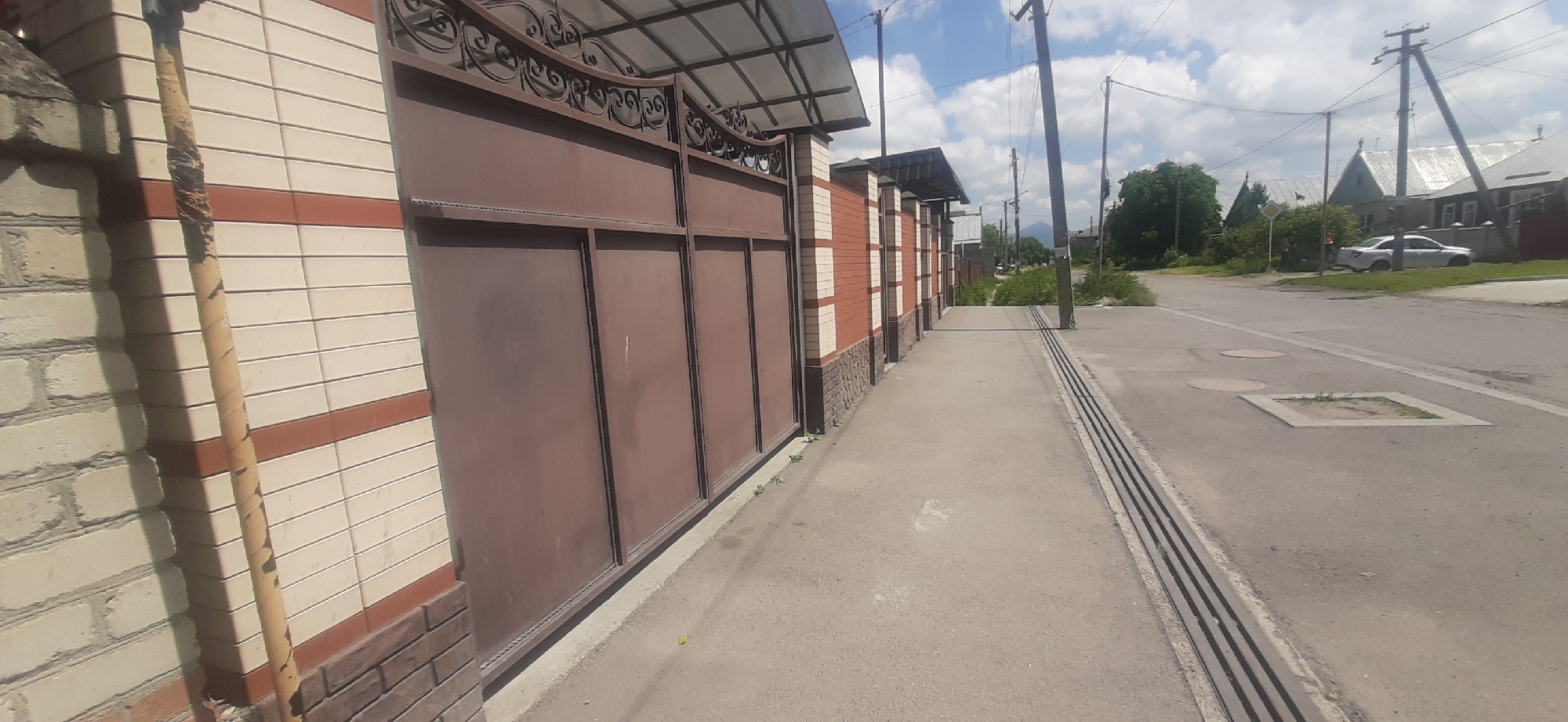 The image shows a street scene in the city of Yessentukskaya, Russia, on a clear, partly cloudy day. In the foreground, a tall, brown metal gate with decorative wrought iron elements at the top stands on the left, adjacent to a brick wall. The wall is constructed with alternating bands of light cream and reddish-brown bricks. A yellow pipe, wrapped with tape in several places, runs vertically along the left side of the wall and in front of the gate.
A concrete sidewalk runs alongside the wall and gate, leading away from the viewer and into the distance. The sidewalk is bordered on the right by a metal drainage grate that runs parallel to the street. The street itself is visible to the right of the sidewalk, with a white car parked on the grass verge on the far side.
In the background, several residential buildings with metal roofs are visible, along with trees and power lines. The overall impression is of a quiet residential street in a rural or semi-urban area.
