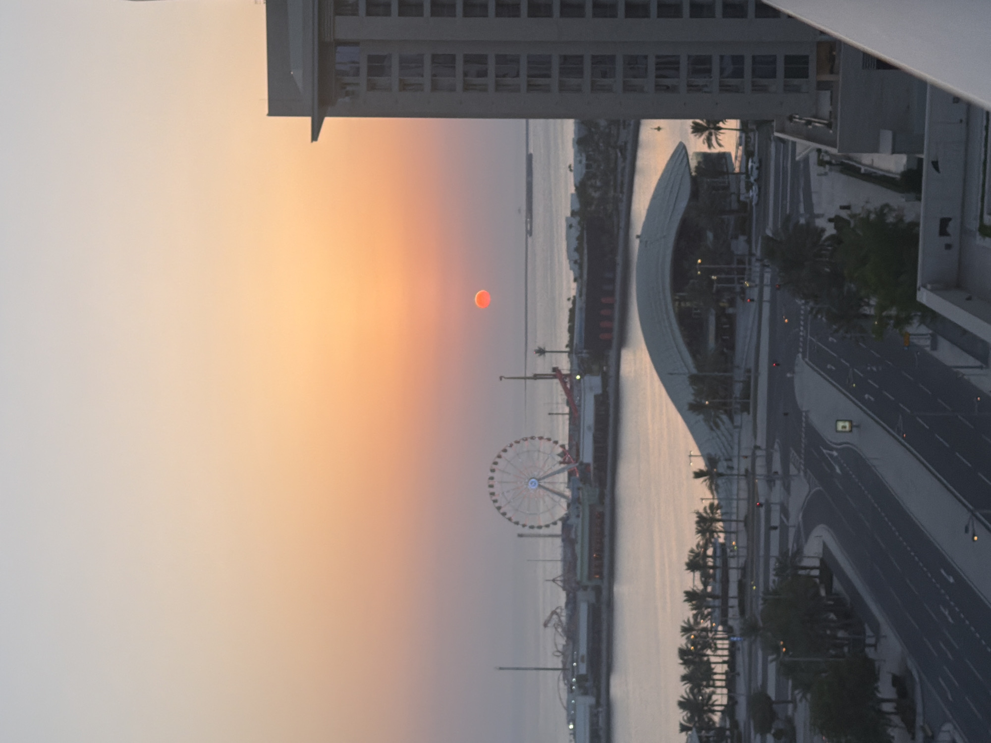 A high-angle, full shot captures a city skyline at sunset. The sun, a small, bright orange orb, hangs in the hazy orange and pastel-colored sky, casting a soft glow over the scene. In the foreground, the right side of the frame is dominated by a tall, modern building with multiple floors and windows. Below, a wide road curves through the cityscape, lined with palm trees and streetlights. Further into the background, a body of water is visible, with a large Ferris wheel and amusement park structures on the opposite shore. A domed building with a curved roof is also present. The overall impression is one of a tranquil urban landscape at dusk.