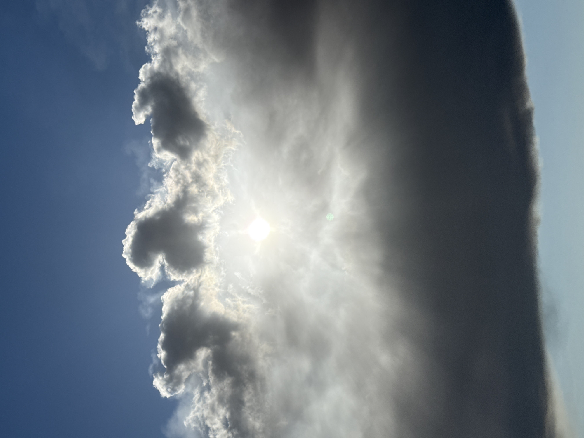 The image captures a dramatic sky scene dominated by clouds and sunlight, oriented vertically. On the left side, a clear, deep blue sky is visible. Toward the center, the bright sun shines intensely, partially obscured by a patch of white and light grey clouds, casting a strong glare and radiating light. To the right, a massive, dark grey cloud formation occupies a significant portion of the frame, appearing dense and almost wall-like. Its edge, particularly on the far right, shows a faint, soft illumination where it meets a lighter, possibly hazy, sky.
Various other clouds are scattered throughout the mid-ground, exhibiting a range of densities and shades from bright white where the sun illuminates them, to darker greys. The lighting suggests daytime. There are no people, man-made objects, or visible text in the photograph. The scene depicts dynamic weather conditions, likely a mix of sun and heavy cloud cover over Perm, Russia, creating a visually striking contrast between light and shadow in the sky.