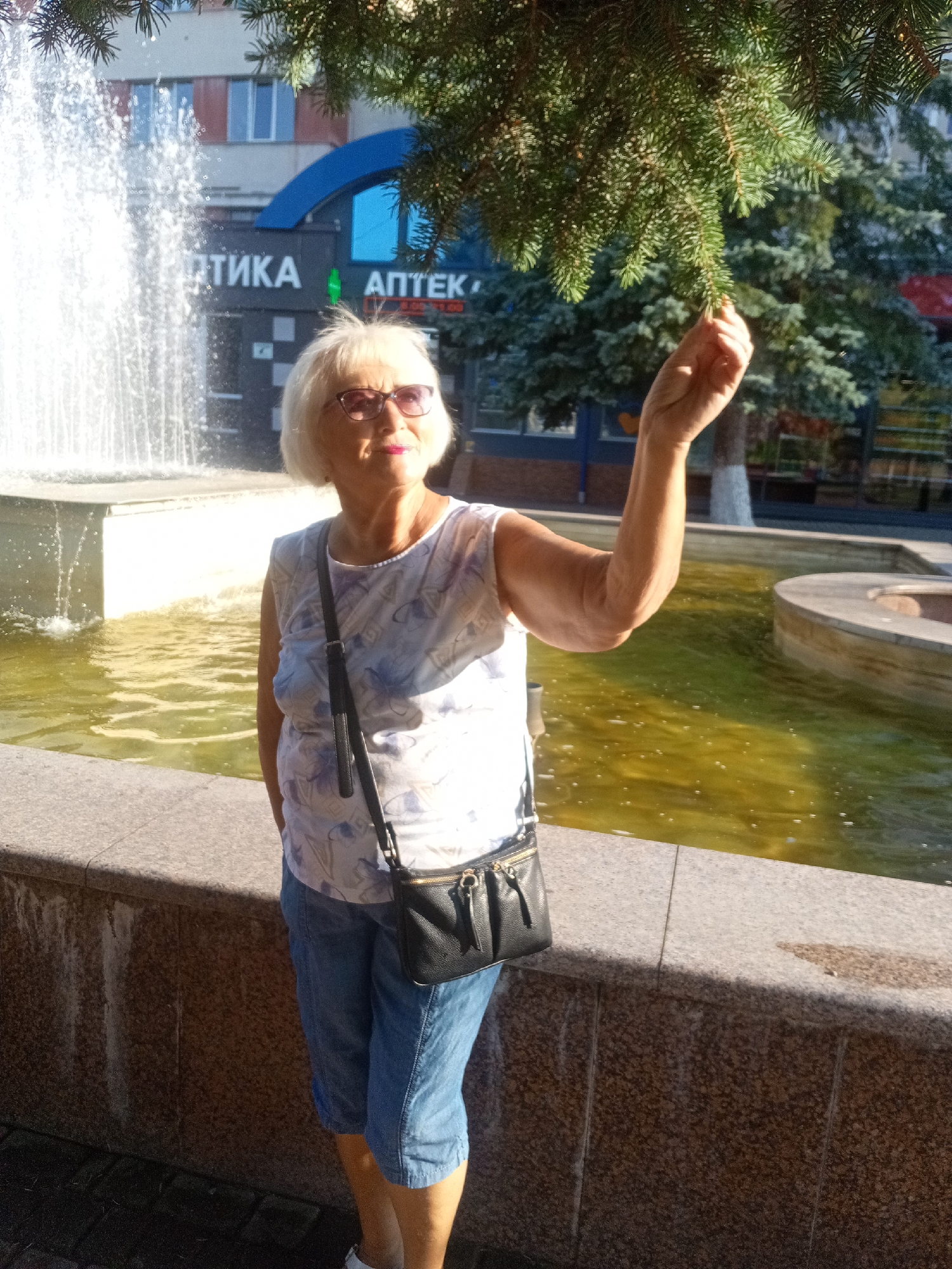 A woman with blonde hair and glasses stands in front of a fountain in Tiachiv, Ukraine. She is wearing a white sleeveless top with a floral pattern and blue denim shorts. A black crossbody bag is slung over her shoulder. Her right arm is raised, and she is touching a tree branch overhead. The fountain water is a murky yellow-green color, and a building with signs in Cyrillic is visible in the background. The sun is shining, casting shadows.