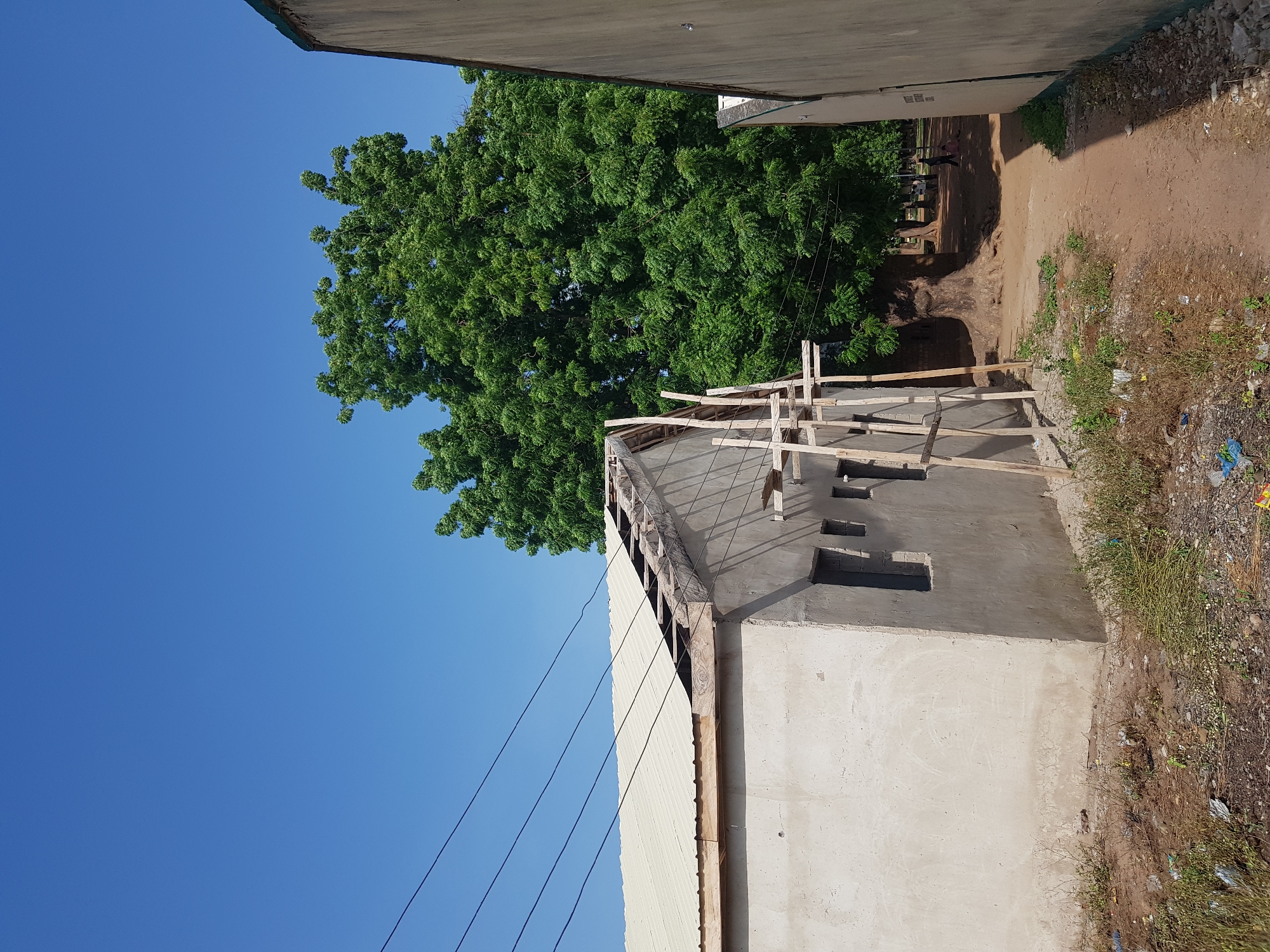 The image depicts an outdoor scene in Jimeta, Nigeria, with a clear blue sky and bright daylight. The primary subjects are a building under construction and a large, leafy green tree. The building is partially finished, with visible scaffolding and exposed wooden beams on its roofline. The walls are plastered, and there are openings that appear to be windows or doorways. The roof of the building is partially covered with corrugated metal sheeting.
In the background, the large tree dominates the left side of the frame, its dense foliage creating a significant canopy. Beyond the tree and construction, a dirt path is visible, with several people walking on it in the distance. Some are standing near the tree, suggesting a communal area or pathway. The ground surrounding the construction site is uneven and appears to be a mix of dirt, gravel, and some dry vegetation. There are also signs of discarded waste, such as plastic bags, visible on the ground near the building.
The overall impression is one of ongoing development and a typical day in a Nigerian town. The lighting suggests it is daytime, likely late morning or afternoon, given the clear sky and the shadows cast. There is no visible text in the image. The scene is neutral, conveying a sense of everyday activity rather than any specific event or strong emotion.