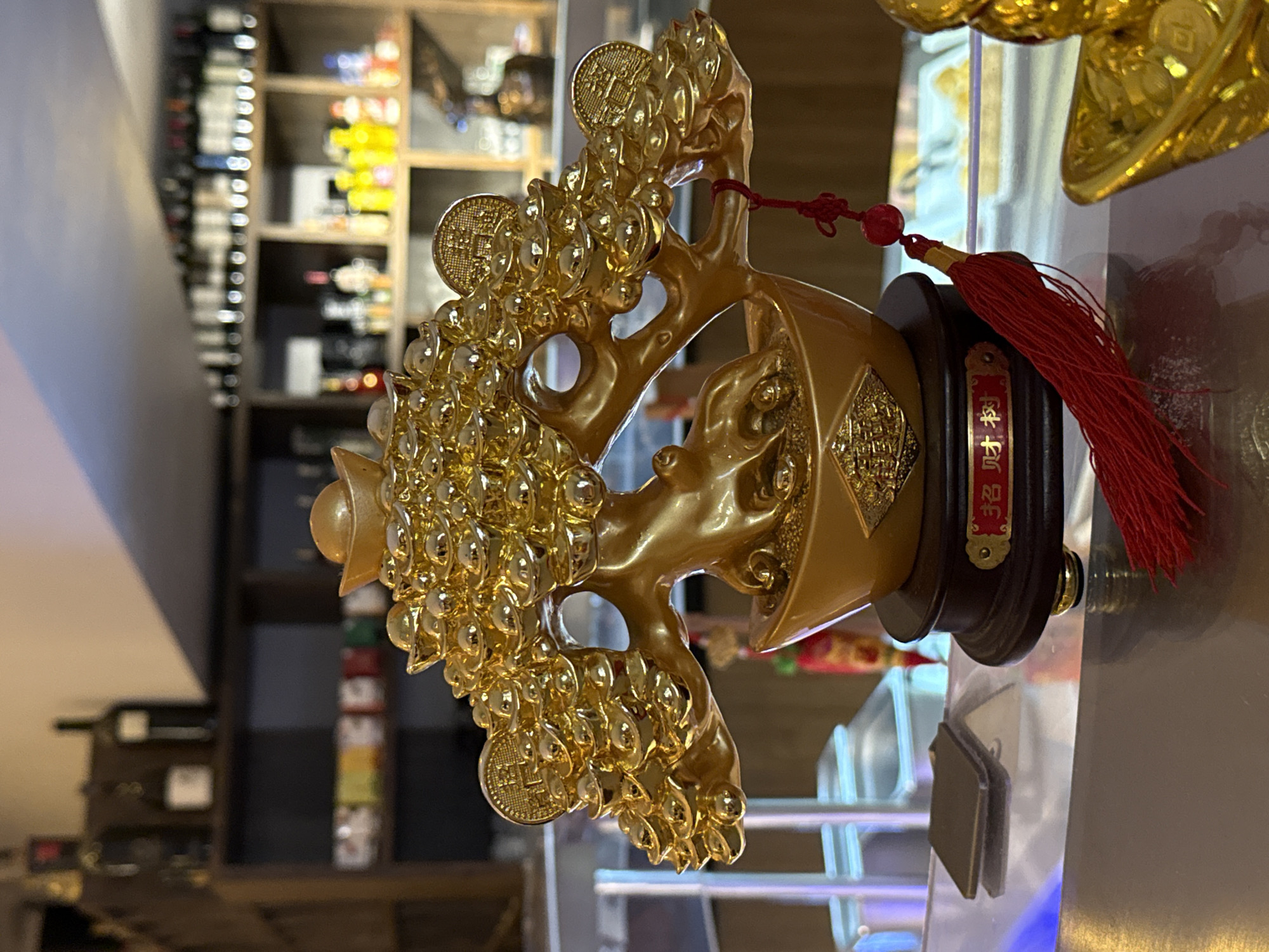 A close-up, eye-level shot features a golden, decorative tree ornament with Chinese inscriptions on its base. The tree is adorned with numerous golden ingots and coins, resembling a wealth tree. A red tassel hangs from one of the branches. The ornament rests on a dark wooden base with gold accents. The background is blurred, showing shelves stocked with bottles of wine and other items, suggesting an indoor setting possibly a bar or restaurant. The overall lighting is somewhat dim with a blueish tint from an unseen light source. The scene is located in Nordhausen, Germany, and appears to be a still life with no human presence or activity.