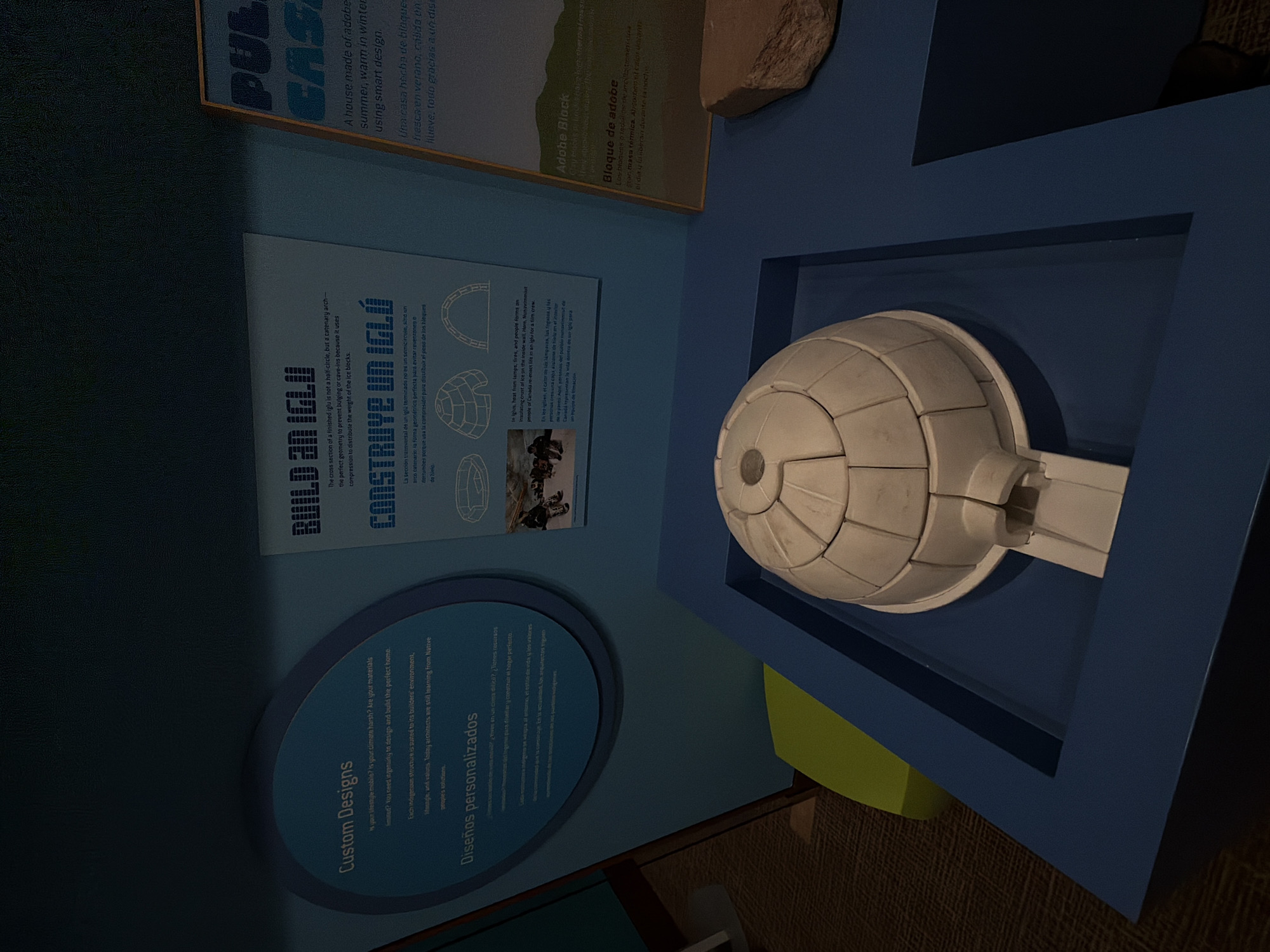 The image depicts an indoor exhibit, likely in a museum, featuring a model of an igloo and informational text. The exhibit is set against a blue backdrop with text in English and Spanish. One panel displays the title "BUILD AN IGLU" and "CONSTRUYE UN IGLÚ," discussing the geometry and construction of igloos, with accompanying diagrams and a photograph of people in an igloo. Another panel, partially visible on the right, has the title "PUBLIC CASTLE" and "BLOQUE DE ADOBE," discussing adobe dwellings. In the foreground, a white, segmented model of an igloo sits on a blue platform. To the right of the igloo model, a rough, brown stone is displayed. The overall environment suggests an educational display about traditional housing structures. No people are visible in the image.
