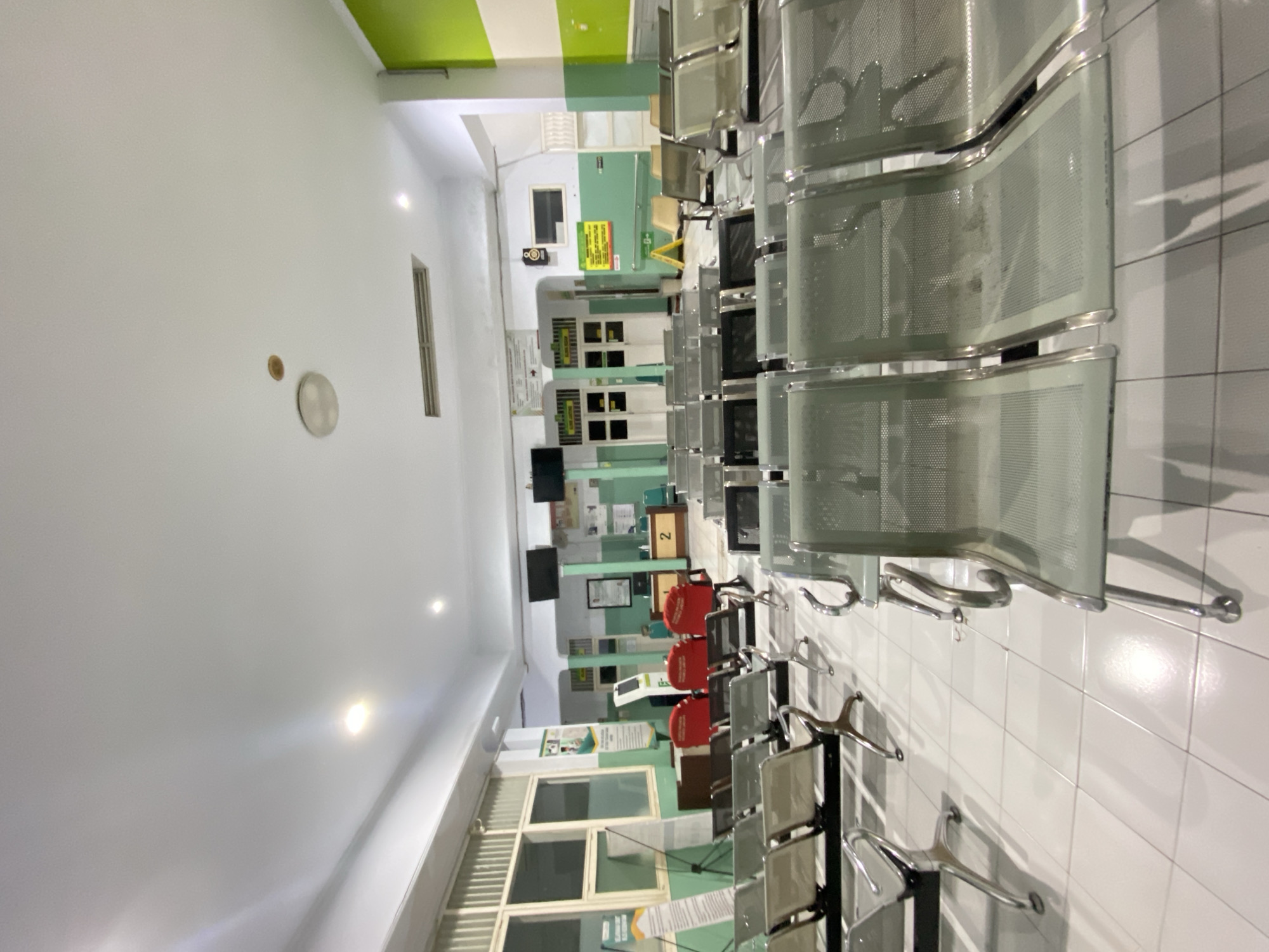 The image depicts a waiting area with rows of empty metal chairs arranged on a tiled floor. The walls are painted in a combination of white and light green. In the background, there are windows with vertical blinds, a reception desk with a sign showing the number "2", and several doorways. There are also two large screens mounted on the wall. The lighting appears to be artificial, with recessed lights in the ceiling. The scene suggests a public space, possibly a clinic or a government office, located in Margoyoso, Indonesia.