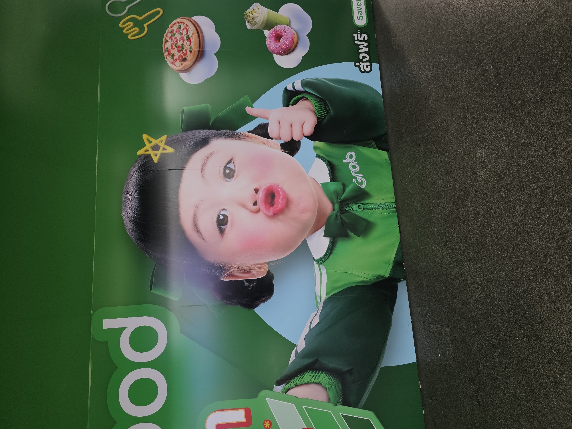 The image is a close-up photograph of a billboard featuring a cartoon-style depiction of a young girl with rosy cheeks and an open mouth, puckering as if to kiss. She has dark hair styled in pigtails with a yellow star clip. The girl is wearing a green uniform with white trim and the "Grab" logo on the chest. She is giving a thumbs-up gesture.
To the right of the girl, there are illustrations of food items: a pizza on a cloud, a pink donut, and a green drink. Above these, stylized yellow cutlery (a spoon and fork) are depicted.
In the lower right corner of the billboard, there is text in Thai which translates to "Free Delivery" and "Saver".
The billboard is predominantly green and blue. The bottom portion of the image shows a dark grey, textured surface that appears to be the ground or a wall at the base of the billboard.
The image was taken in Bangkok, Thailand, likely in an outdoor urban setting where such advertisements are common. The billboard is mounted on a wall. The lighting suggests it's daytime, with no harsh shadows indicating direct sunlight. The overall impression is an advertisement for Grab, a popular ride-hailing and delivery service in Southeast Asia.