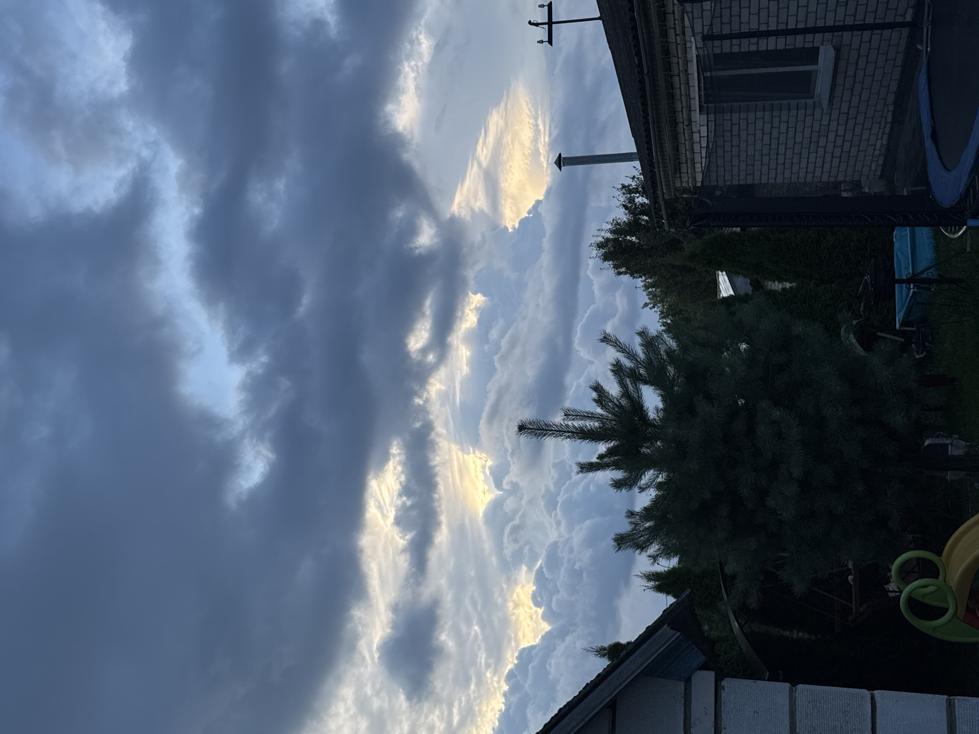 The image shows a suburban backyard in Maladzyechna, Belarus, with a dramatic sky filled with dark, heavy clouds that are partially illuminated by the setting sun. In the foreground, the roofline of a brick house and a smaller, light-colored building are visible, along with the dark silhouette of a coniferous tree. A trampoline and a small blue pool are also present in the yard. The overall mood is somber, with the impending storm creating a sense of foreboding.