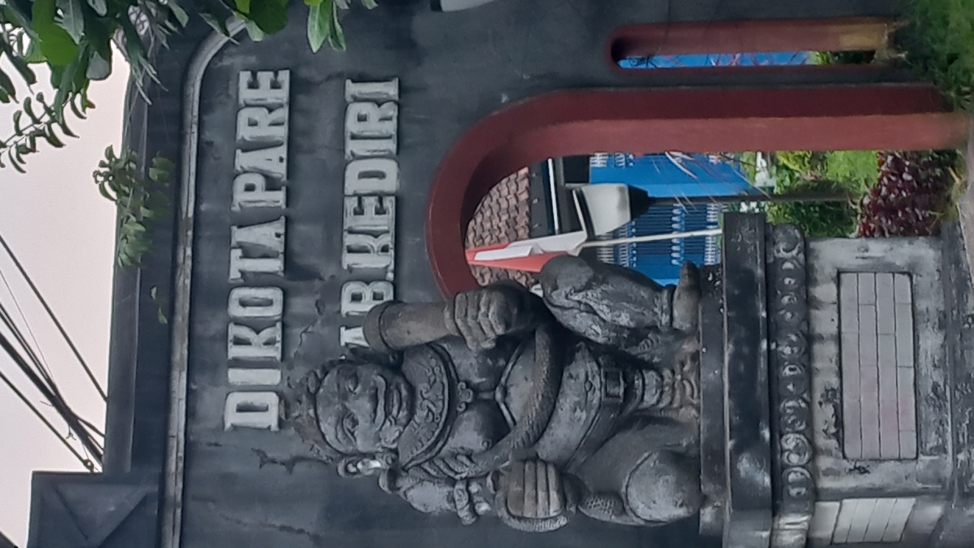 The image shows a stone statue of a mythical figure in front of a gate with lettering. The statue is dark gray and depicts a crouching, muscular figure with a round face, prominent ears, a mustache, and a headdress. It holds a club-like object in its right hand and what appears to be a smaller object in its left. The statue is seated on a tiered pedestal. Above and behind the statue, the gate features raised, white lettering that reads "DIKOTA PARE" on the upper line and "AB KEDIRI" on the lower line. The gate itself is partially visible, with a red archway framing the view beyond. Through the archway, a blue building with white bars is visible, along with a flagpole flying the Indonesian flag. Green foliage and some red plants are also present in the foreground and around the gate. The overall scene suggests an entrance to a location in Pare, Kediri, Indonesia, possibly a cultural site or administrative building. The sky above is overcast.