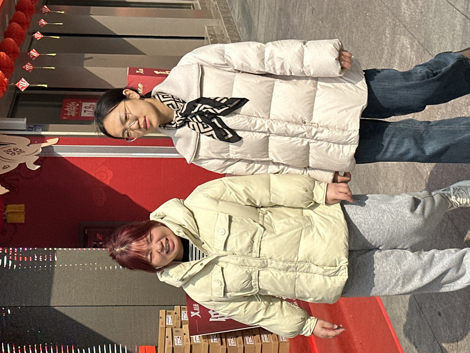 Two women stand outside in front of a red wall with Chinese decorations. The woman on top wears a white puffer jacket, jeans, glasses, and a black and white scarf. The woman below wears a light yellow puffer jacket, grey sweatpants, and has reddish-purple hair. They both face forward, smiling slightly. The background includes a building, pavement, and what appears to be boxes.