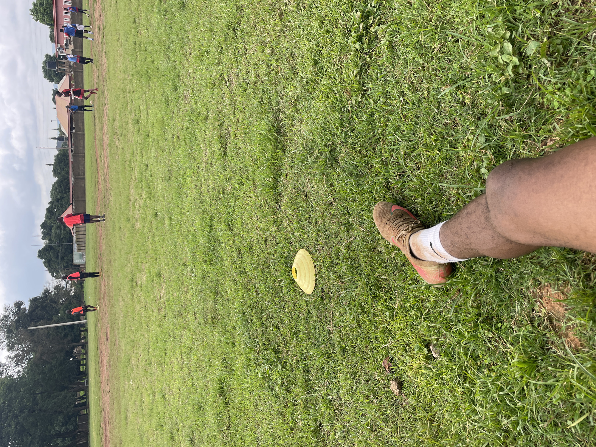 The image depicts an outdoor scene on a grassy field, likely a football pitch. In the foreground, a person's leg and foot are visible, wearing a football boot and a white sock. The boot is brown and appears to be muddy. A yellow training cone is placed on the grass to the left of the leg. In the mid-ground and background, several individuals are engaged in what appears to be a football training session or game. They are scattered across the field, some wearing bright orange vests, others in red and blue. The environment is characterized by lush green grass, trees along the perimeter, and buildings in the distance, including structures that suggest urban development. The sky is overcast with clouds. The presence of training cones and multiple individuals playing football strongly indicates a sports facility within the city of Jos, Nigeria.