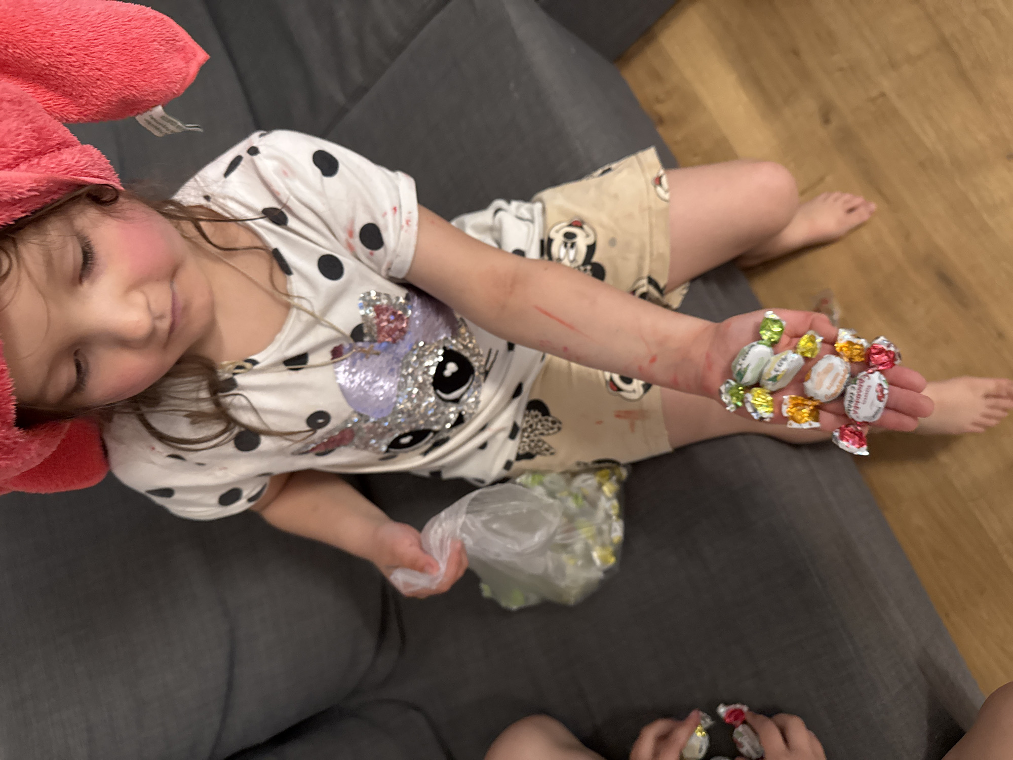 A young child with a pink towel on their head is seated on a gray couch, holding a handful of wrapped candies in their right hand. A plastic bag containing more candies is in their left hand. They are wearing a white t-shirt with black polka dots and an image of a cartoon character. The child's shorts have cartoon characters printed on them. The setting appears to be indoors with a wooden floor visible in the background. Another child's legs and hands are seen at the bottom of the picture with two candies on the floor. There is no information to confirm the location is Koltushi, Russia.