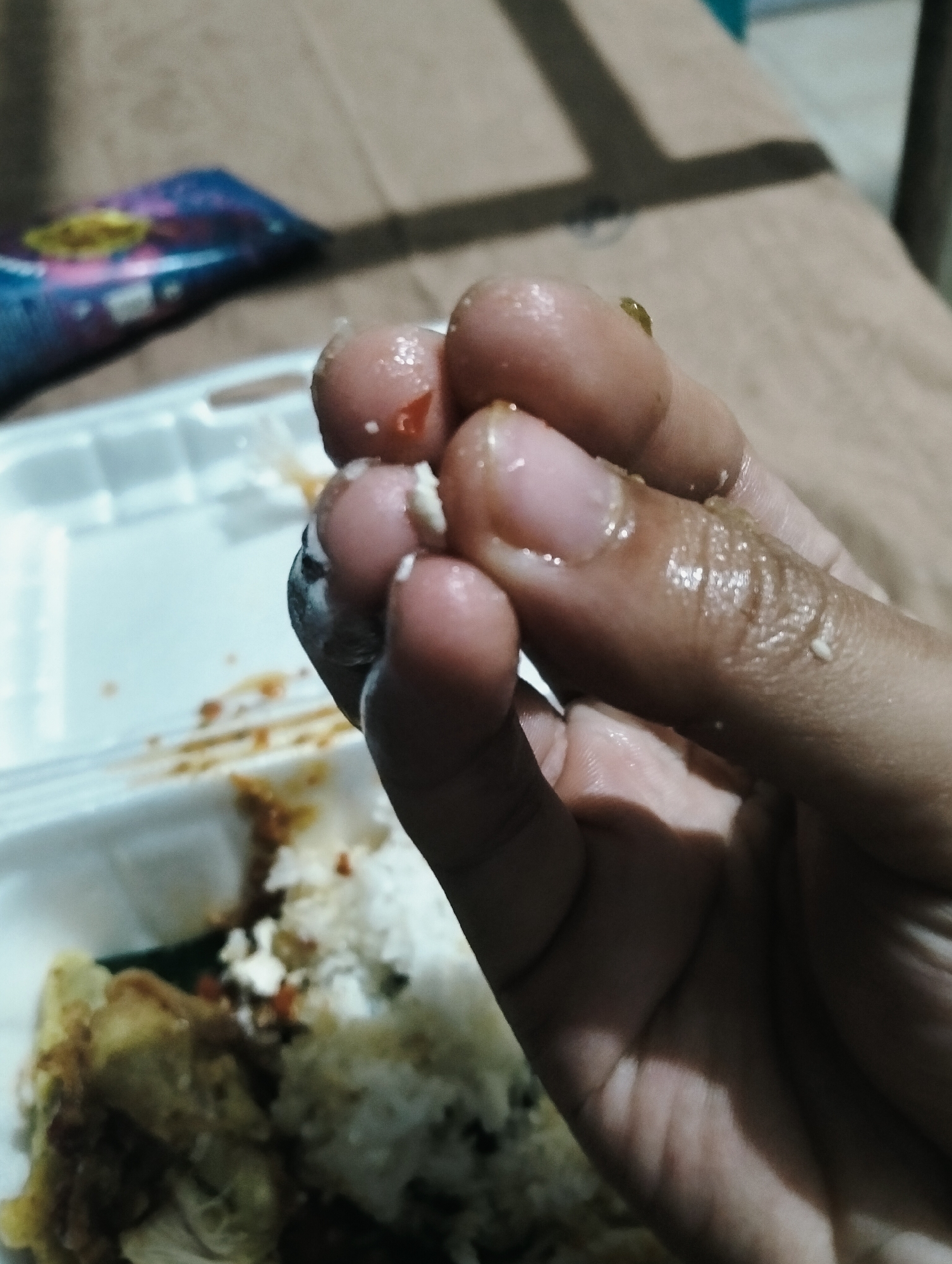 A close-up shot shows a person's hand, with fingers soiled with food remnants. The hand is positioned over a white styrofoam container holding rice and other food items. The background is out of focus, but appears to be a surface with some shadows, possibly a table or bed. A blurred colorful package is visible in the upper left corner. The overall impression is of a casual, possibly home-like setting. The image does not provide any specific information about the city of Medan, Indonesia.