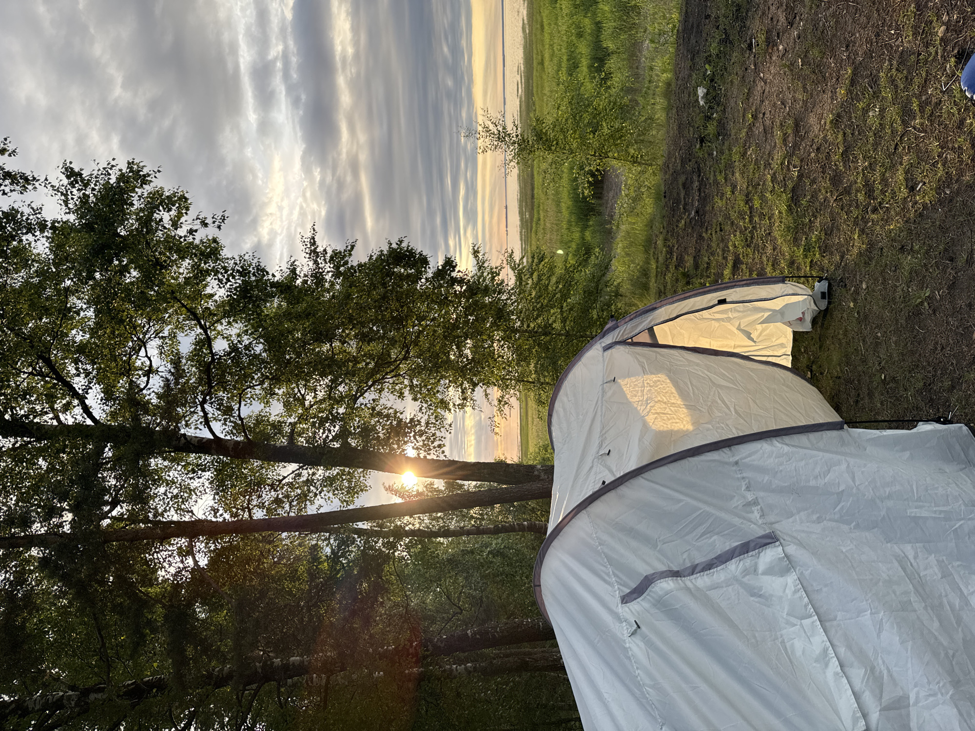 A white tent is pitched on a patch of grass and dirt, with its entrance open and illuminated from within. Trees with green foliage are visible in the foreground and on the left, with sunlight filtering through them. In the background, a body of water stretches to the horizon under a cloudy sky. The scene appears to be a campsite, possibly located in Bolshaya Izhora, Russia, given the natural landscape and proximity to water. There are no people visible in the image.