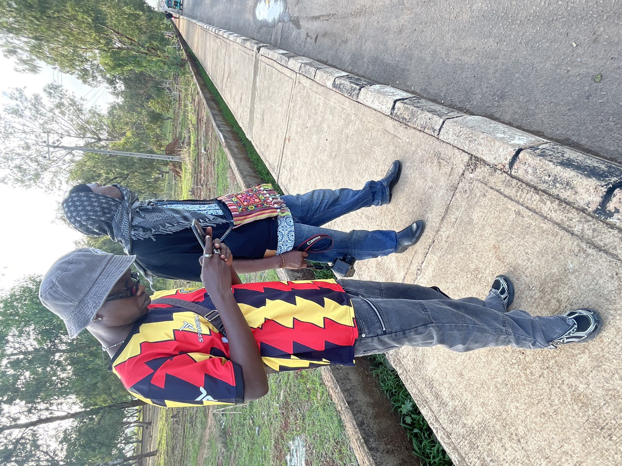 Two African men are standing on a concrete sidewalk adjacent to a paved road. The man closest to the camera is wearing a red, yellow, and black shirt, grey jeans, black and white sneakers, and a grey bucket hat. He is holding a smartphone and has a messenger bag slung across his torso. The second man is wearing blue jeans, black shoes, a black scarf, and is holding a Canon camera. The scene is in a grassy area with trees in the background. The location is Bar Jirgi Summa, Nigeria.