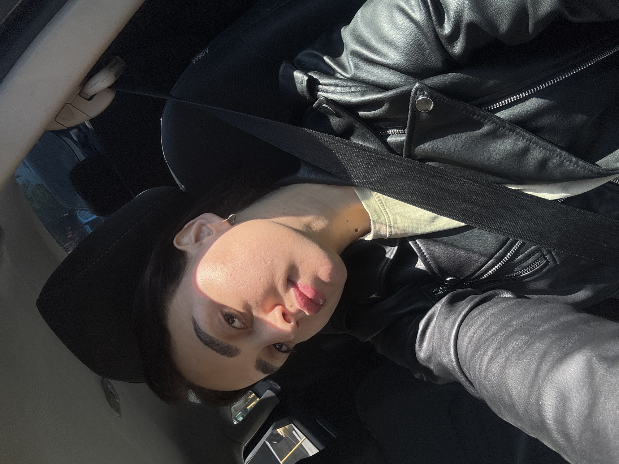A person with dark hair wearing a black hat and black leather jacket sits in a car with a seatbelt fastened. The person's face is visible, showing dark eyebrows and lips. The interior of the car is black, and sunlight shines on the person's face. The location is not discernible.