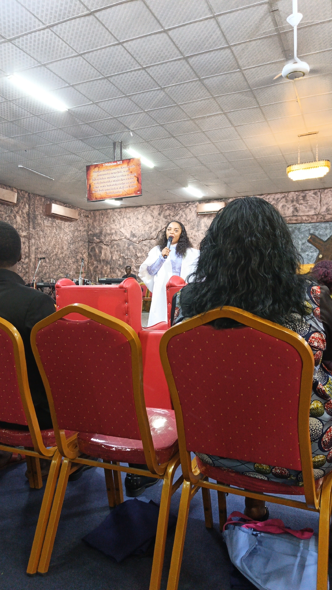 In the interior of a church, a woman in a white robe and blue scarf stands on a raised platform, speaking into a microphone. She has dark, curly hair and is looking towards the audience. In front of the platform are several rows of red chairs with gold frames, some of which are occupied by members of the congregation. The church walls have a textured, stone-like appearance, and there is a large sign hanging from the ceiling with text in a language other than English. The lighting suggests it is daytime. The scene appears to be within a religious gathering.