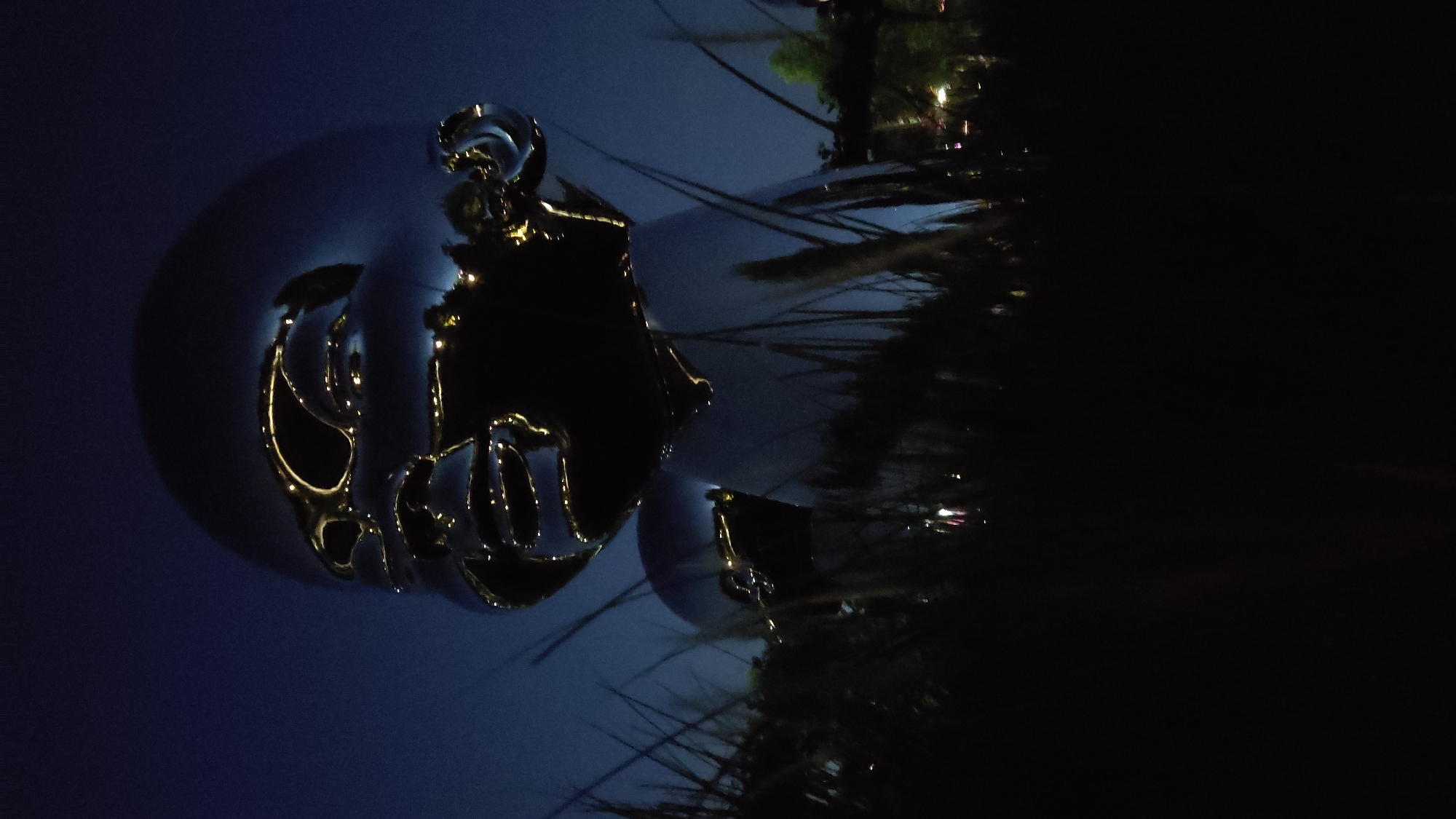 The image captures a nighttime outdoor scene featuring a large, reflective spherical sculpture, likely a public art installation. The sphere itself is a deep blue or purple hue, and its highly polished surface reflects subtle elements of the surroundings. Adorning the sphere is an intricate, golden metallic design, which appears to be a stylized, abstract face or emblem, possibly incorporating elements reminiscent of a character like Mickey Mouse due to prominent rounded shapes. The golden outlines of this design shine brightly, illuminated against the darker sphere.
In the background, dark, dense foliage, possibly trees or bushes, is silhouetted against the night sky, occupying the right side of the frame. Scattered faint lights, suggesting artificial illumination from distant buildings or streetlights, are visible amidst and beyond the foliage. These distant lights and the dark natural elements are also subtly reflected in the glossy surface of the main sculpture.
The scene is tranquil and artistic, characteristic of a public space or park illuminated after dark. The time of day is clearly night, with the dominant color palette being dark blues, purples, and black, punctuated by the gleaming golden accents of the sculpture. There is no visible text or human activity, emphasizing the sculptural elements. This art piece is located in Krasnodar, Russia.