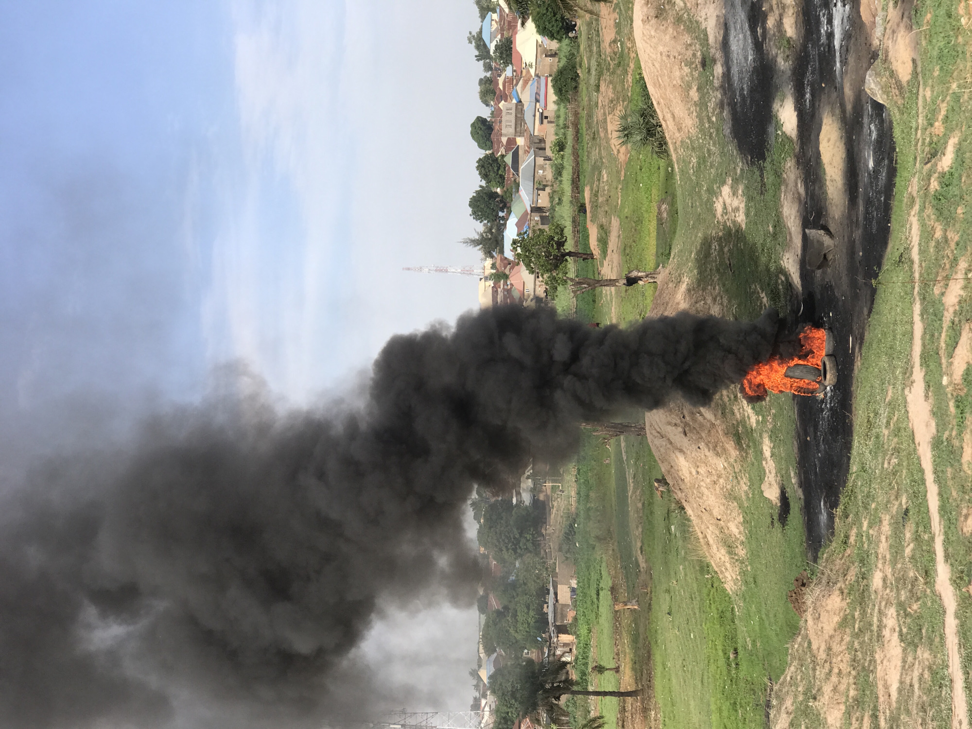 The image, taken in Abuja, Nigeria, captures a significant fire producing a massive plume of thick, black smoke against a daytime sky. Intense orange flames are visible at the source of the fire, originating from an object burning on the ground.
The fire is located in an open, semi-rural or peri-urban landscape. The immediate area around the blaze consists of patches of green grass, bare earth, and prominent light-colored rock formations or mounds. Some of these rocky surfaces bear dark, charred stains, hinting at prior burning activities at this spot. A dirt path or track is visible winding through the grassy terrain.
A dense column of black smoke rises steadily and expands across the sky, indicative of incomplete combustion, possibly from materials such as tires or plastic waste. Above the smoke, the sky is light blue with scattered white clouds, confirming it is daytime with seemingly calm weather.
In the background, beyond the open ground and rock formations, a clustered settlement or village is visible. Numerous buildings with roofs of various colors, including red, blue, and grey, indicate a residential area. Scattered trees and other vegetation are present throughout the scene, particularly near the settlement. A tall, slender communication tower or antenna stands in the distance.
No people are explicitly visible in the frame, and there is no discernible text. The image documents an active burning event, likely related to waste disposal, with the heavy smoke representing a notable environmental impact.