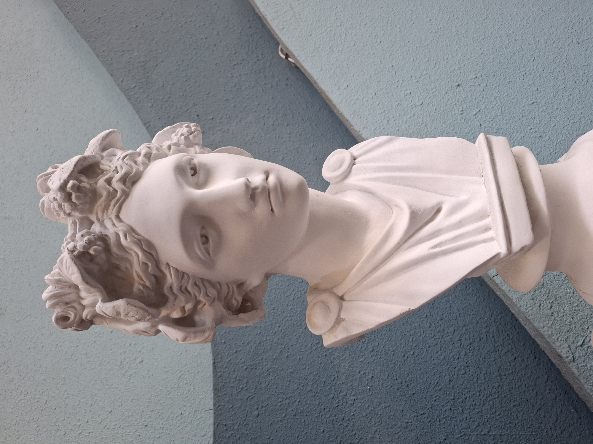 A white plaster bust of a woman with ornate hair is positioned in front of a blue, textured wall. The wall has a diagonal, lighter gray line intersecting it. The bust is a close-up shot, focusing on the head, neck, and upper chest. The woman's hair is elaborately styled with what appears to be leaves or flowers. She wears a garment draped over her shoulders with circular embellishments. The lighting casts subtle shadows, highlighting the sculpted features of the bust. There is no indication of the city of Nha Trang Ward, Vietnam, in this image.