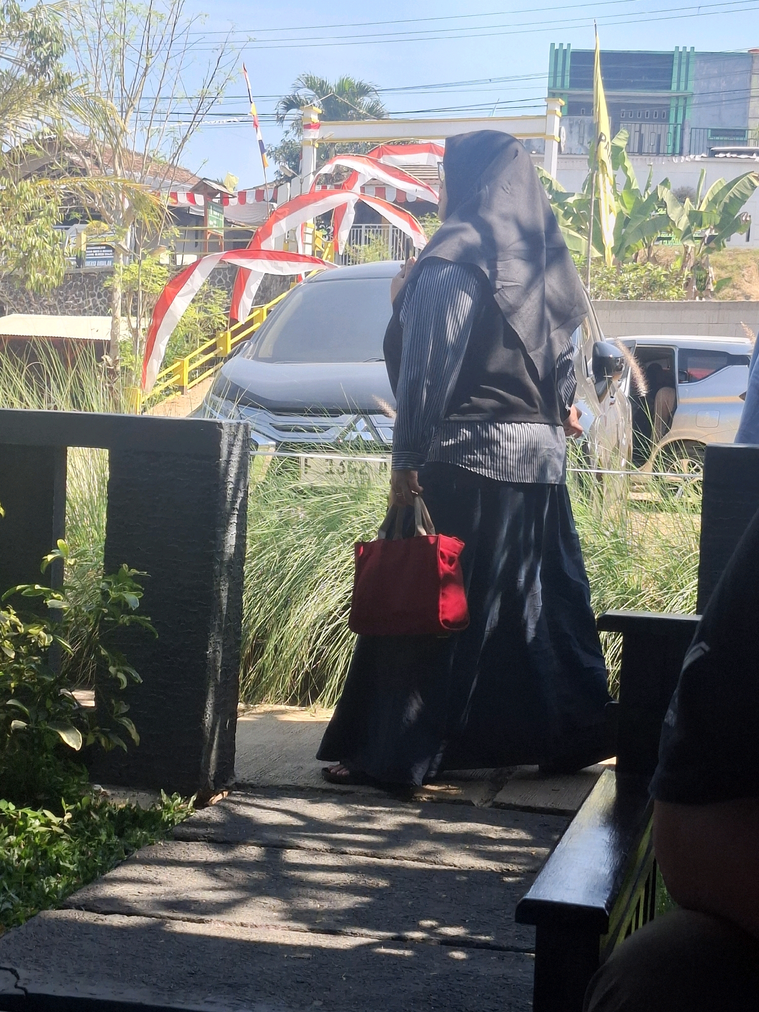 A side-view, medium shot captures a person wearing a black hijab, a striped long-sleeved shirt, and a dark skirt, standing on a paved walkway with shadows from overhead foliage. The person is holding a red handbag with beige straps in their right hand. To the left of the person, a dark gray, textured pillar is visible, with lush green plants at its base and to its left. In the background, a dark-colored SUV is parked, and beyond that, a light-colored car with its door open. Further in the background, there is a building with green accents, yellow poles, and festive red and white arched decorations. Tall green grass and trees are abundant throughout the scene. The image appears to be taken outdoors, suggesting a public space in Cibentang, Indonesia, possibly during a celebration due to the decorations.