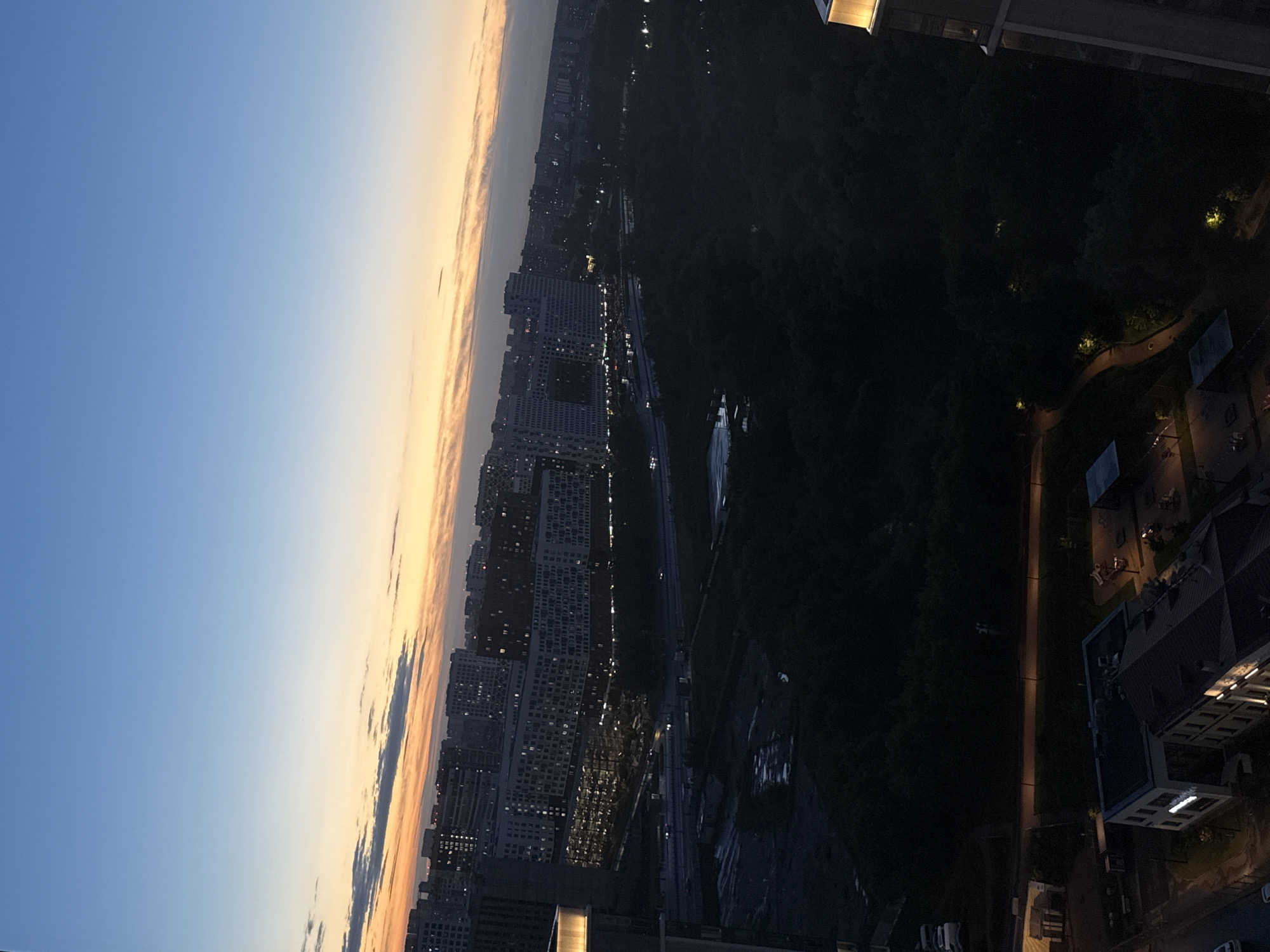 The image depicts an aerial view of the city of Dolgoprudny, Russia, at dusk. The sky transitions from a deep blue at the top to hues of orange and yellow near the horizon, with wispy clouds. Below the sky, a sprawling cityscape is visible, characterized by numerous multi-story buildings with illuminated windows, indicating that it is evening. A dark, dense forest occupies a significant portion of the middle ground, separating the main urban area from the foreground. In the foreground, more buildings are visible, some with artificial lighting, and a few vehicles can be spotted on the roads. The overall scene conveys a sense of urban development adjacent to a natural landscape, observed during the twilight hours.