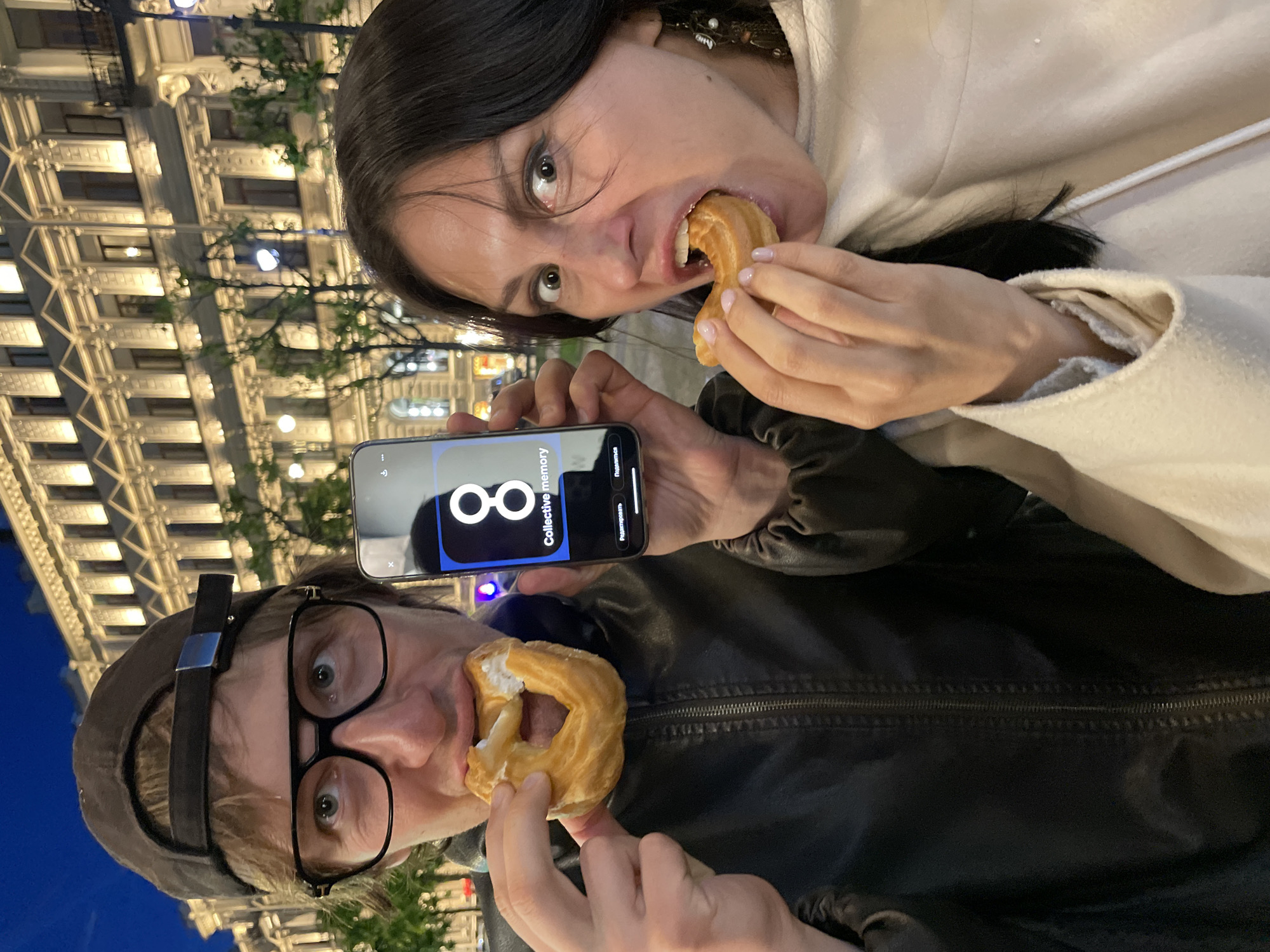 In this medium shot, captured at dusk in Saint Petersburg, Russia, a man and a woman are the central figures. The man, wearing a dark baseball cap and glasses, is in the foreground on the left, biting into a circular pastry. He is dressed in a black leather jacket. Beside him, on the right, the woman, with dark hair and an expressive face, is also taking a bite from a similar pastry. She is wearing a light-colored coat. The woman holds a smartphone in her right hand, displaying the application "Collective memory" with a stylized number 8. Behind them, a grand, illuminated building with multiple windows and architectural details forms the backdrop, suggesting a cityscape at night. Trees and streetlights are visible in the background, further contributing to the urban evening setting. The overall scene depicts two individuals enjoying a snack outdoors, with the city's architecture providing a striking backdrop.