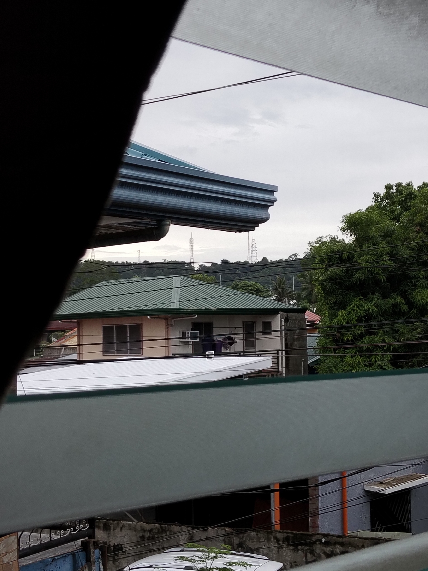 The image shows a view of a residential area in Davao City, Philippines, likely from an elevated position. The foreground is dominated by horizontal blinds or louvers, with a dark, angled object on the left obscuring part of the view.
In the midground, a two-story house with a green corrugated roof is visible. It has cream-colored walls with windows covered by screens. To the right of this house, there is a large, leafy green tree. Electrical wires crisscross the scene, obscuring parts of the background.
Further in the background, rolling hills covered in trees are visible, with what appear to be telecommunication towers on the crests. The sky is overcast, suggesting a cloudy day. The overall impression is of a typical urban or suburban landscape in a tropical climate.