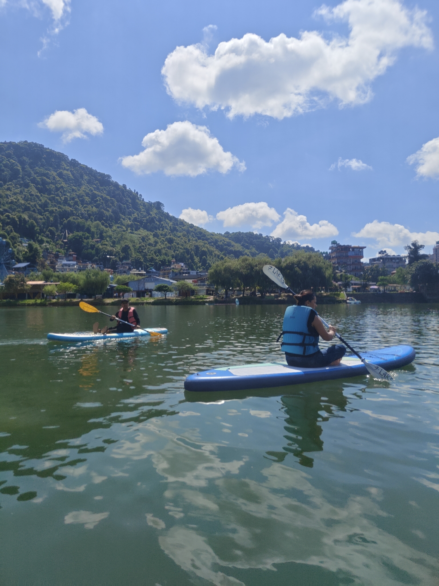 In Pokhara, Nepal, two individuals are paddleboarding on Fewa Lake under a bright blue sky dotted with fluffy white clouds. A lush green hillside, densely covered with trees and featuring scattered buildings, forms the backdrop. The lake's surface is calm and reflects the sky and surrounding scenery. In the foreground, a person wearing a blue life vest and dark pants is seated on a paddleboard, actively paddling with a double-bladed oar. To the left and slightly further away, another person is also paddleboarding on a lighter-colored board, using a single-bladed oar. The overall scene depicts a serene day of outdoor recreation on the famous lake.