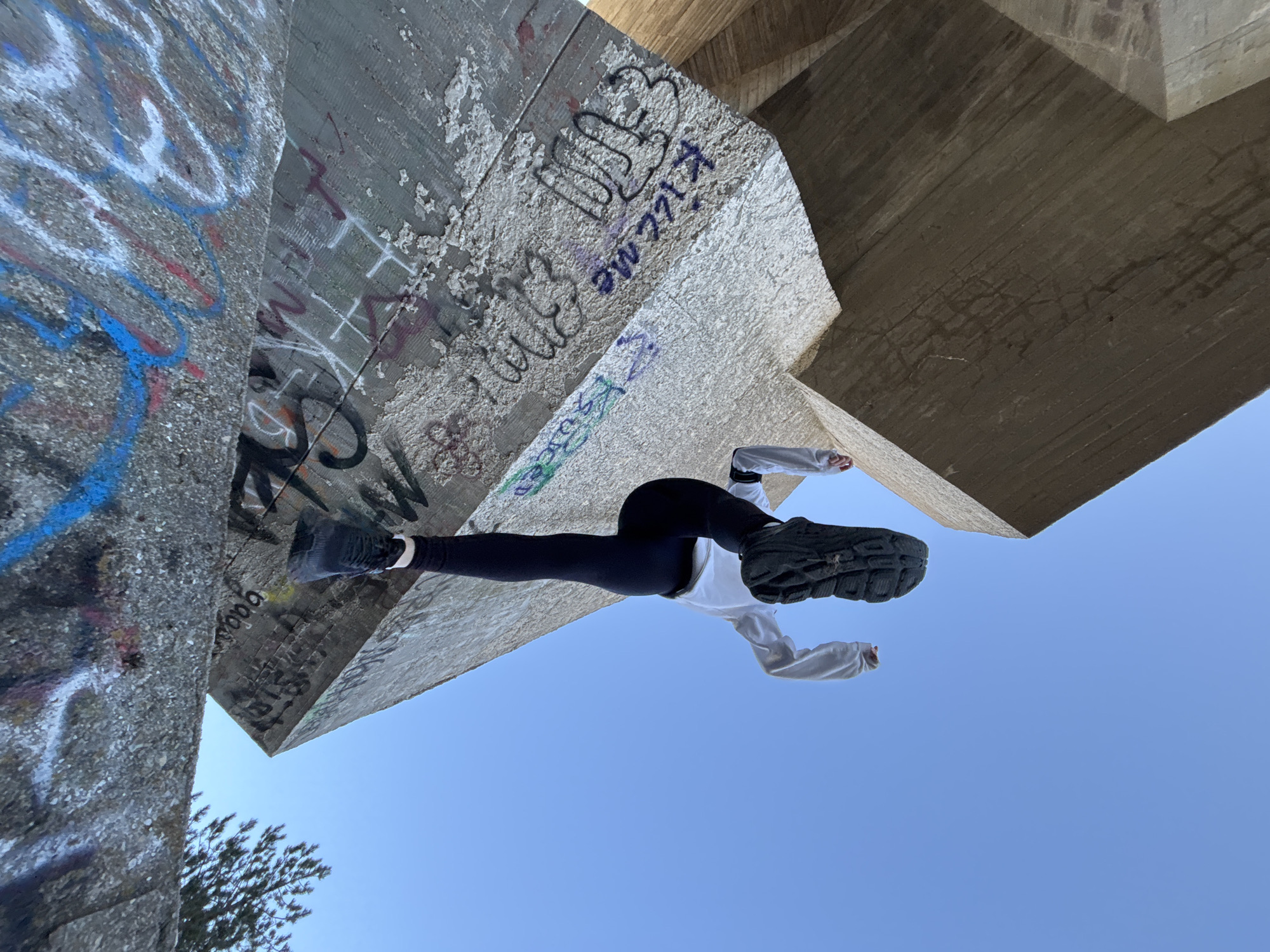 A dynamic, low-angle shot captures a person positioned against a massive, graffiti-covered concrete structure, likely an overpass or large architectural element in Varna, Bulgaria. The person appears suspended or in an acrobatic pose, with their body oriented horizontally in the frame, but seemingly upside down relative to the concrete blocks they interact with. They wear black leggings, a white long-sleeved top, and dark athletic shoes with thick, textured soles.
The concrete surfaces are rough, textured, and extensively covered in various graffiti tags and drawings in colors including blue, white, red, purple, and black. Notable visible text among the graffiti includes "Kiss me" written in purple/blue script. The structures themselves show signs of weathering and minor erosion.
The majority of the background is a clear, bright blue sky, indicating a sunny day. A small section of green foliage from a tree is visible in the bottom-left corner, behind one of the concrete blocks. The unique perspective creates an illusion of the person defying gravity, highlighting a playful or performative interaction with the urban environment.