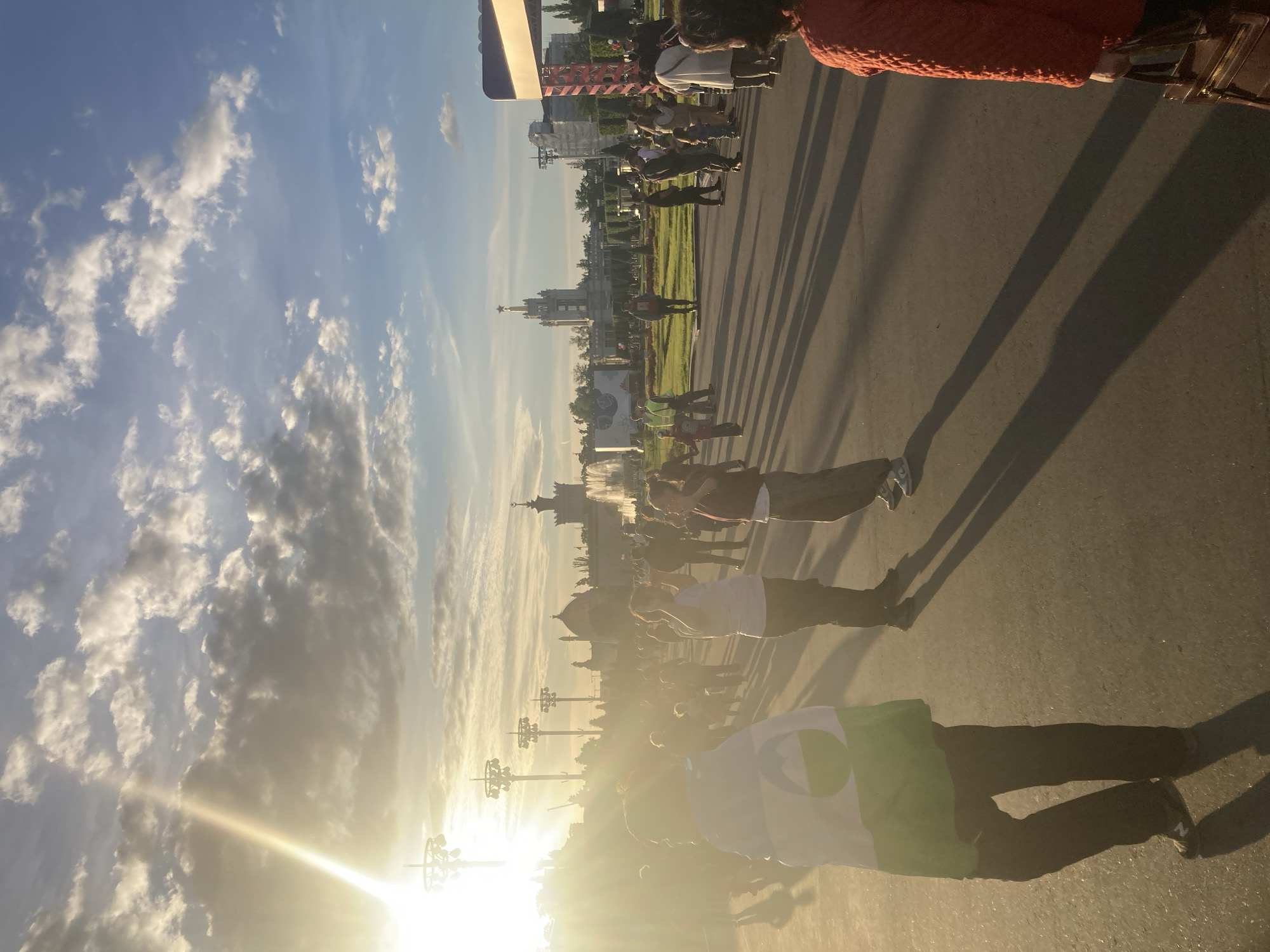 The image depicts an outdoor public gathering in Moscow, Russia, bathed in the warm, golden light of a late afternoon or early evening. The sun, low on the right side of the frame, casts a strong lens flare and creates long, distinct shadows stretching across the paved ground towards the left.
Numerous people are present, populating a wide street or plaza. In the immediate foreground, a person is visible holding a green and white flag featuring a dark circular design. Other individuals are scattered throughout the middle ground, some appearing to walk or stand, all casting prominent shadows. The overall atmosphere suggests a public event, possibly a parade, festival, or demonstration.
In the background, a distinct urban landscape unfolds. Prominent buildings with classical and Soviet-era architecture are visible, including a large structure topped with a spire and a star, characteristic of Moscow's Stalinist architecture. A strip of green lawn separates the paved area from these buildings. Several decorative lampposts or streetlights are silhouetted against the bright sky. The sky above is a clear blue, adorned with scattered white cumulus clouds. No legible text is visible on any banners or signs.