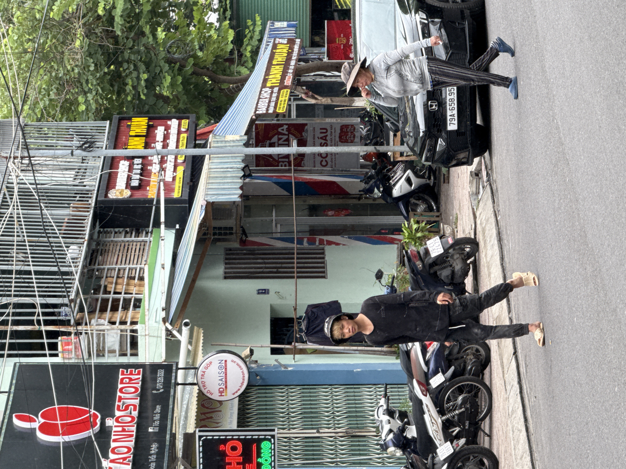 The image shows a street scene in Nha Trang, Vietnam. A young man wearing a black long-sleeved shirt, black pants, and a bucket hat walks across the street towards the right of the frame. He is barefoot except for light-colored sandals. Behind him, parked motorcycles are visible along the sidewalk. On the right side of the street, a woman wearing a grey hooded jacket and striped pants walks across the street towards the left. She is wearing a wide-brimmed hat and appears to be touching her face. A black sedan is parked on the right side of the street. Buildings with signs are visible behind the pedestrians, including a sign for "TAO NHOSSTORE" with a red apple logo, and other signs in Vietnamese. The street is paved, and there is a sidewalk with some greenery. Overhead wires and streetlights are also visible.