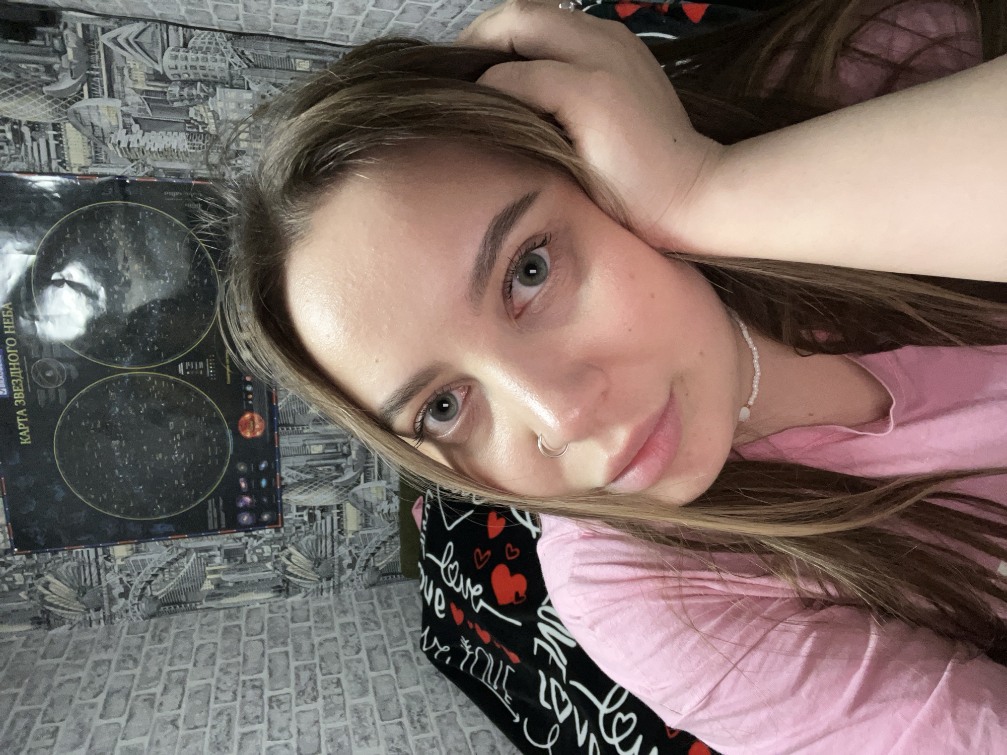 A young woman with fair skin and long brown hair lies on a bed. She wears a pink t-shirt and a beaded necklace. Her right hand is near her head. She has a nose ring. The bed has a black blanket with red hearts and white text. A poster of a star chart is on the wall. The wallpaper is grey and white. The location of the photo is not discernible in the image. The city of Novopokrovskaya, Russia, is not verifiable from this image.