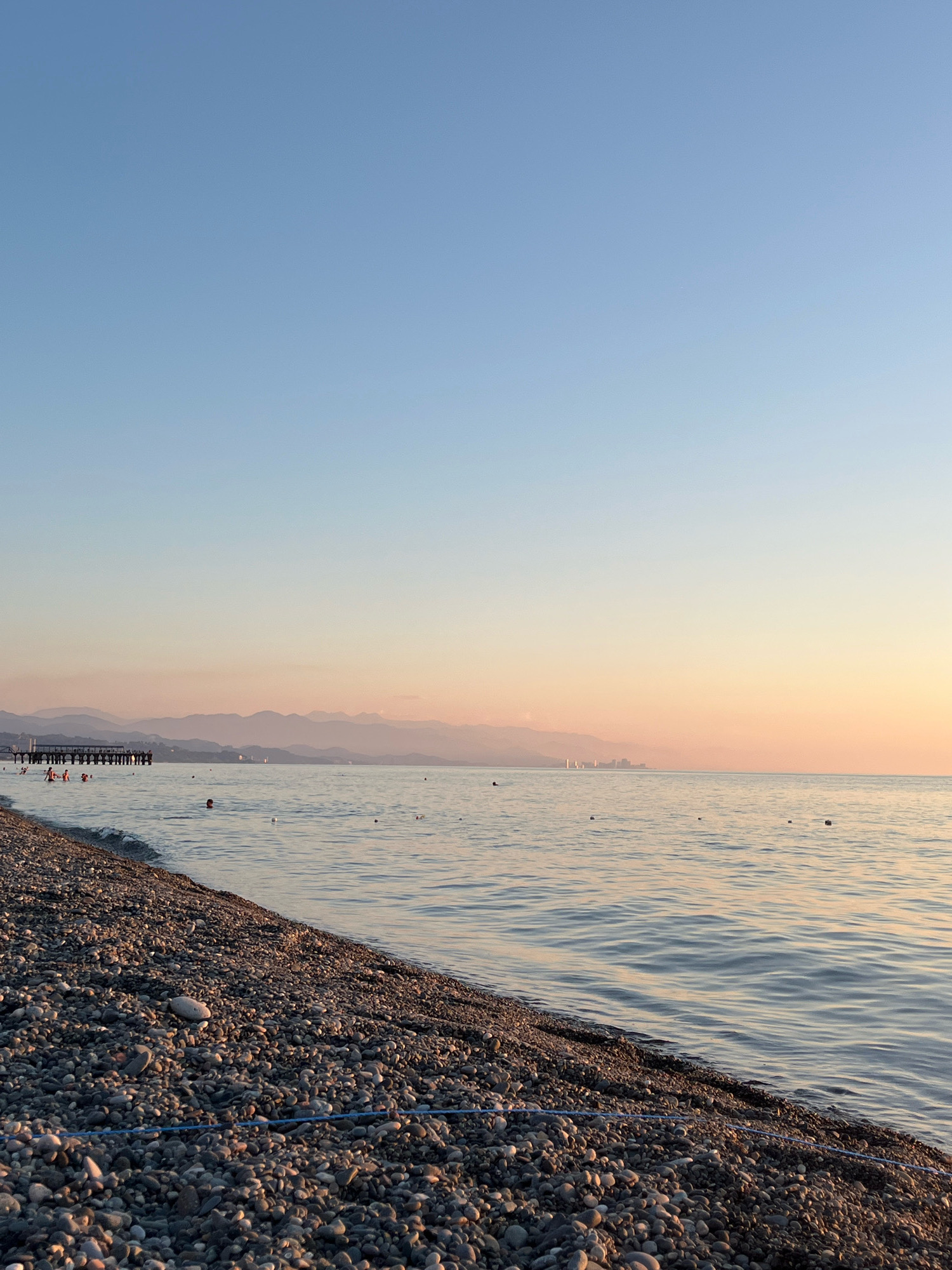 This is a photograph of the Black Sea coast in Kobuleti, Georgia, during sunset. The foreground features a pebble beach with a blue rope laid out horizontally across it. The sea is calm with gentle ripples, reflecting the soft colors of the sky. In the middle ground, several people are visible in the water, some swimming and others standing. A pier extends into the sea from the left side of the frame, with people standing on it. In the distance, a hazy mountain range is silhouetted against the horizon. The sky is a gradient of clear blue at the top, transitioning to soft hues of pink and orange near the horizon, indicative of a sunset.