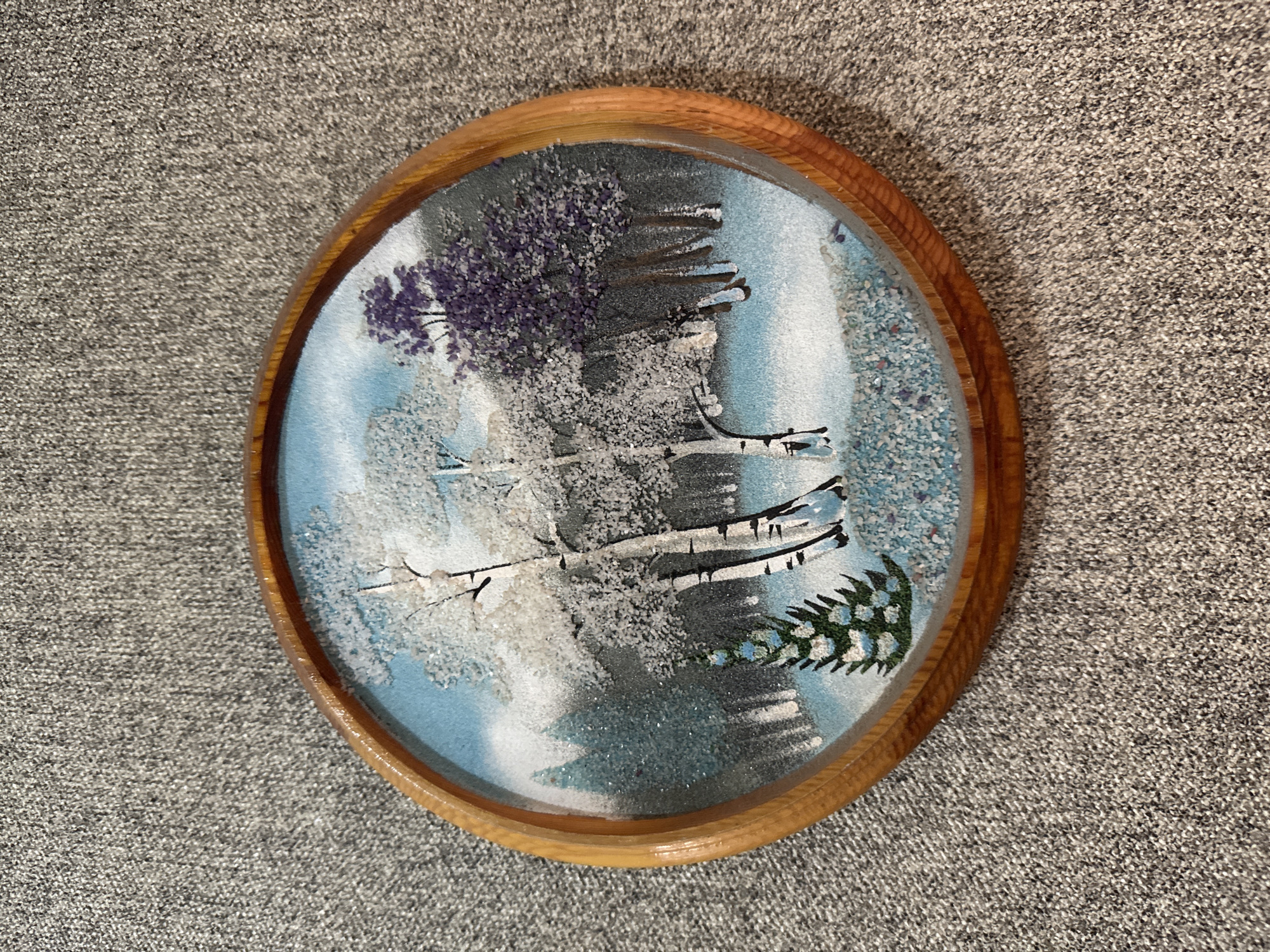 The image shows a small, oval, framed artwork depicting a winter forest scene. The artwork is made of sand or fine glitter and is set against a light blue sky. In the foreground, a small evergreen tree is visible, coated in white material resembling snow. Behind the evergreen, several birch trees with white bark and black markings stand prominently. To the right of the birch trees, a cluster of trees with purple foliage is depicted. The background shows more dark trees and a hazy, snowy landscape. The artwork is encased in a light-colored wooden frame with a ribbed texture. The framed artwork is resting on a grey, textured surface, possibly a carpet or fabric. There are no people or animals in the image. The location in Bishkek City, Kyrgyzstan cannot be determined from the provided image.