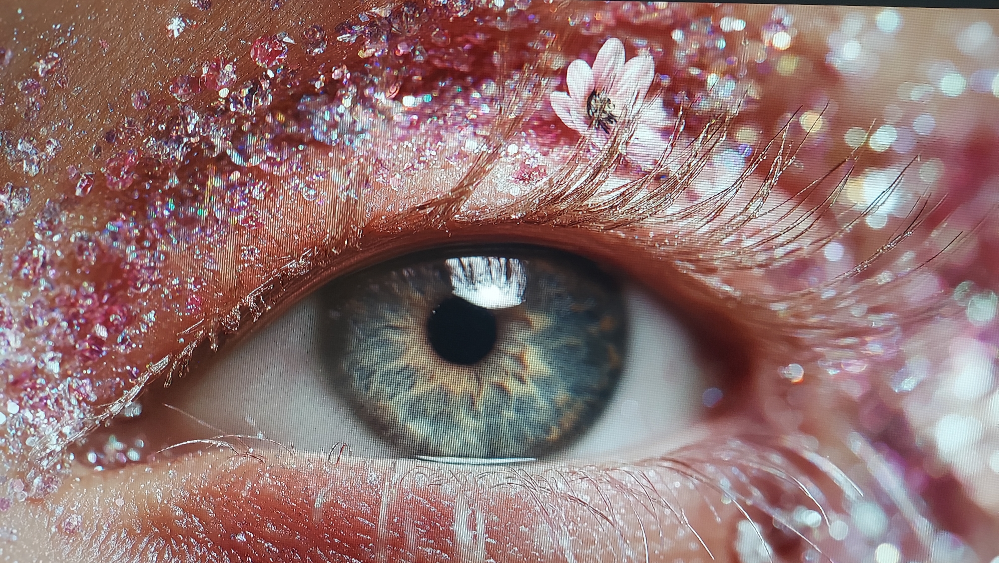 The image is a close-up, macro shot of a person's eye, adorned with pink glitter and a small, delicate white flower. The iris of the eye is a complex pattern of blue and brown hues, and the eyelashes are long and dark, some dusted with glitter. The skin around the eye is textured and appears to have more glitter applied. The background is blurred and bokeh, with soft pink and white lights, suggesting a studio or controlled lighting environment. There is no indication of the city of Tbilisi, Georgia, or any urban environment in this image.