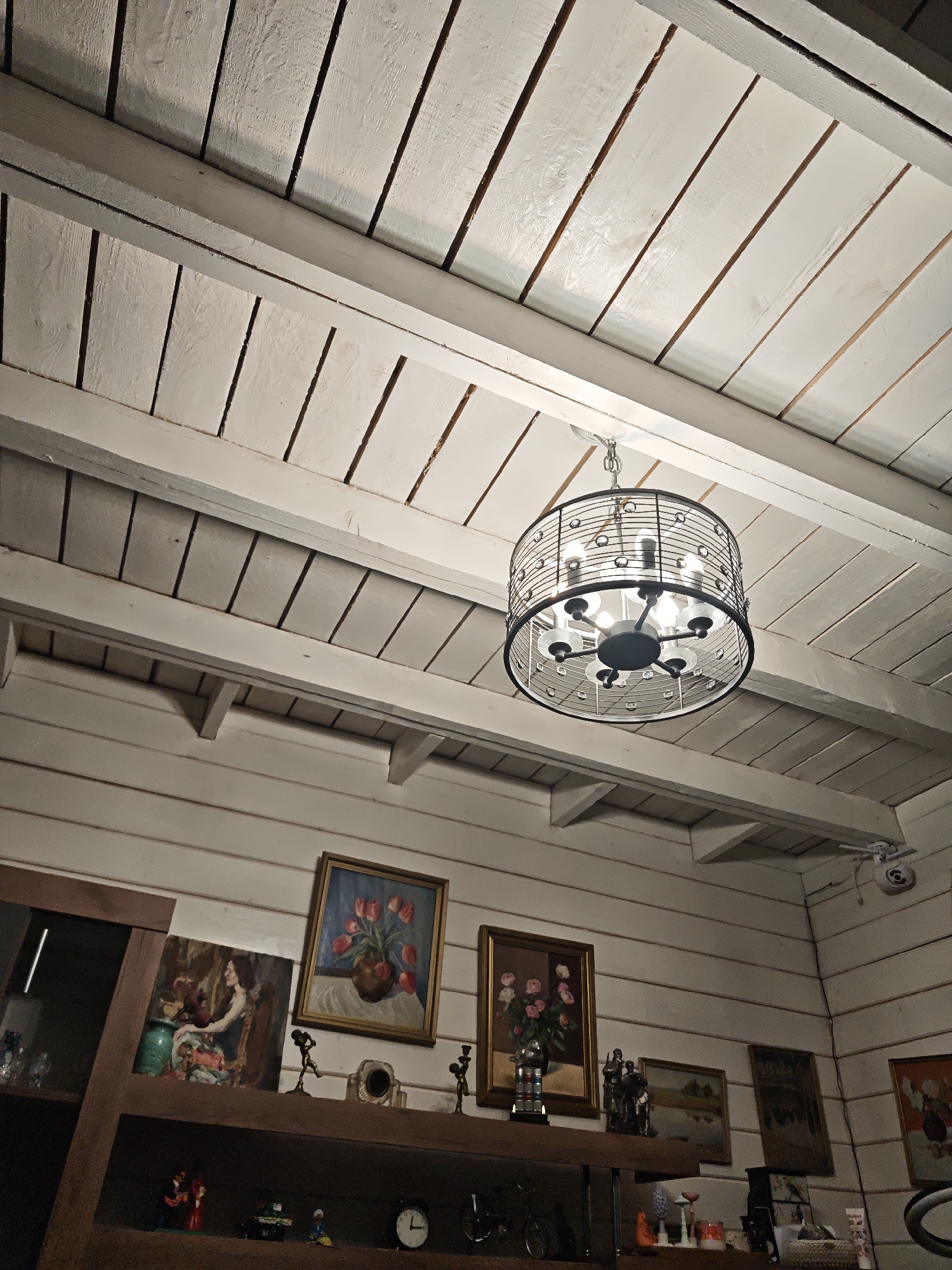 The image shows the interior of a room in Свистуха (кузяевский С/о), Russia, featuring a white wooden paneled ceiling with visible beams and a black, circular chandelier with multiple light bulbs hanging from it. Below the ceiling is a wall adorned with various framed paintings and a dark wooden shelf displaying an assortment of objects, including figurines, decorative items, a clock, and other miscellaneous items. A partial view of a dark wooden cabinet is visible on the left side of the frame. A security camera is visible in the upper right corner of the frame. The room appears well-lit, with the light source primarily coming from the chandelier.