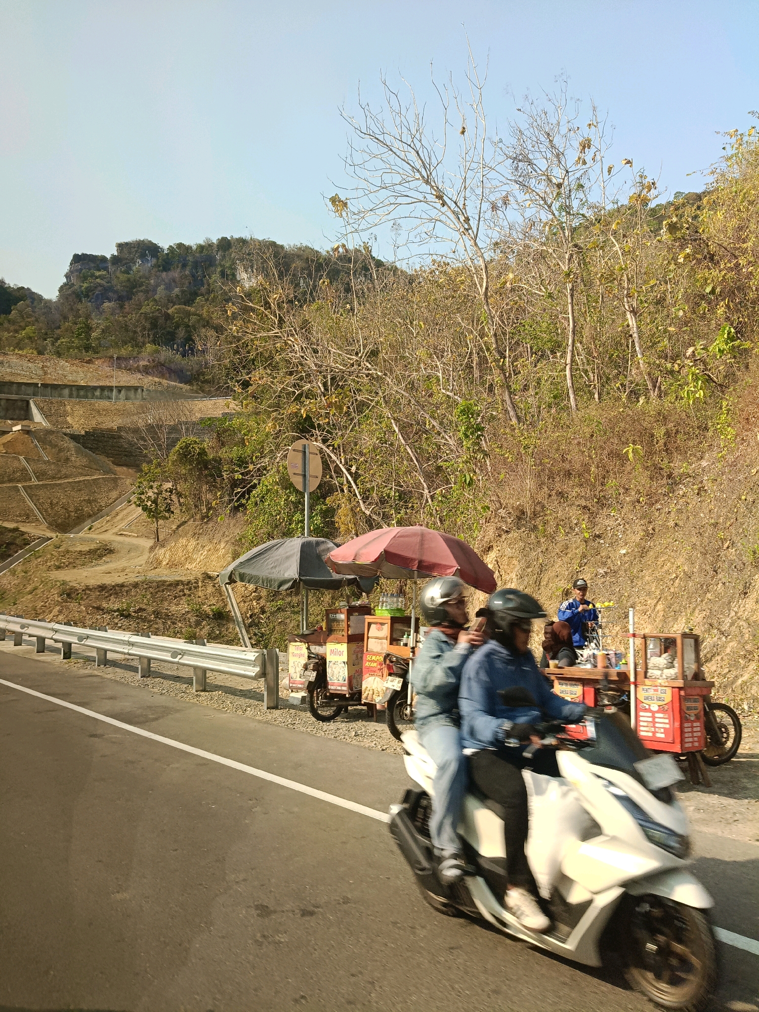 The image depicts a roadside scene near Parangtritis, Indonesia, characterized by a dry, hilly landscape under a clear sky. A paved road runs diagonally from the lower left to the upper right of the frame. On the right side of the road, two food stalls are set up on the side of the embankment. One stall, on the left, has a dark umbrella, and the other, on the right, has a red umbrella. Both stalls are colorful, with visible signage and display cases containing food items. A man in a blue jacket stands behind the stall on the right, attending to customers or preparing food.
In the foreground, a white scooter with two occupants is captured in motion, blurring the details of the riders and the vehicle. The rider in the front wears a blue jacket, and the passenger behind wears a similar jacket. Both are wearing helmets.
Behind the food stalls, the terrain rises into a steep, dry hill covered with sparse vegetation, predominantly trees with bare branches, suggesting a dry season. Terraced slopes are visible on the left side of the road, with retaining walls and pathways. A metal guardrail separates the road from the embankment where the stalls are located. The overall atmosphere is bright and sunny, typical of a tropical environment during dry weather.