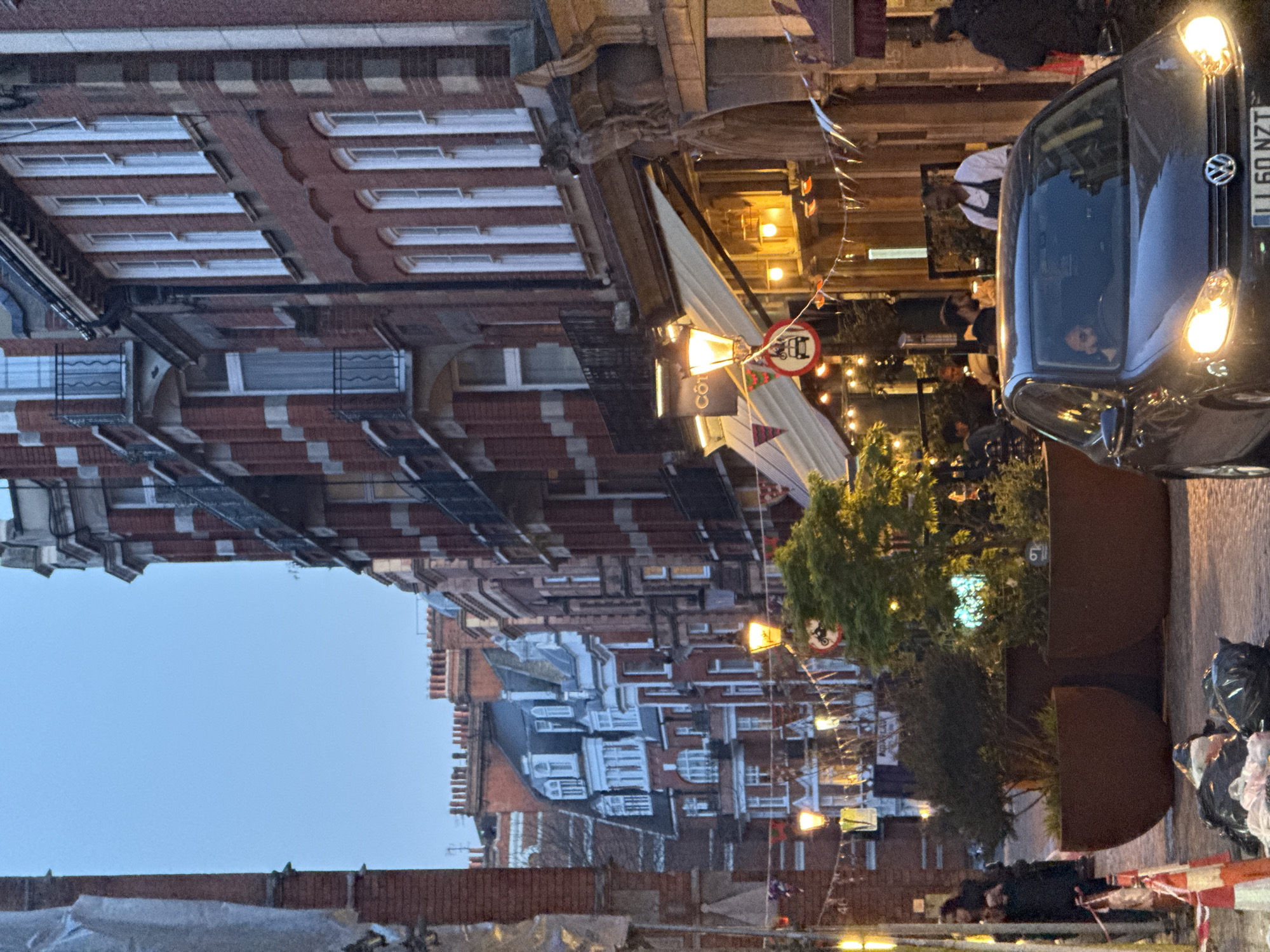 The image depicts a street scene in London, United Kingdom. A dark-colored car is positioned on the right side of the frame, with its headlights illuminated. A man is visible in the driver's seat.
On the left side of the street, there are rows of brick buildings with numerous windows. A restaurant, "CÔT", with an awning is visible. People are seated at outdoor tables enjoying food and drink. There is a motorcycle sign nearby.
Banners are strung across the street. Towards the bottom of the frame, there are rubbish bags, a barrier, and a person is lying on the ground. The sky is visible above the buildings.