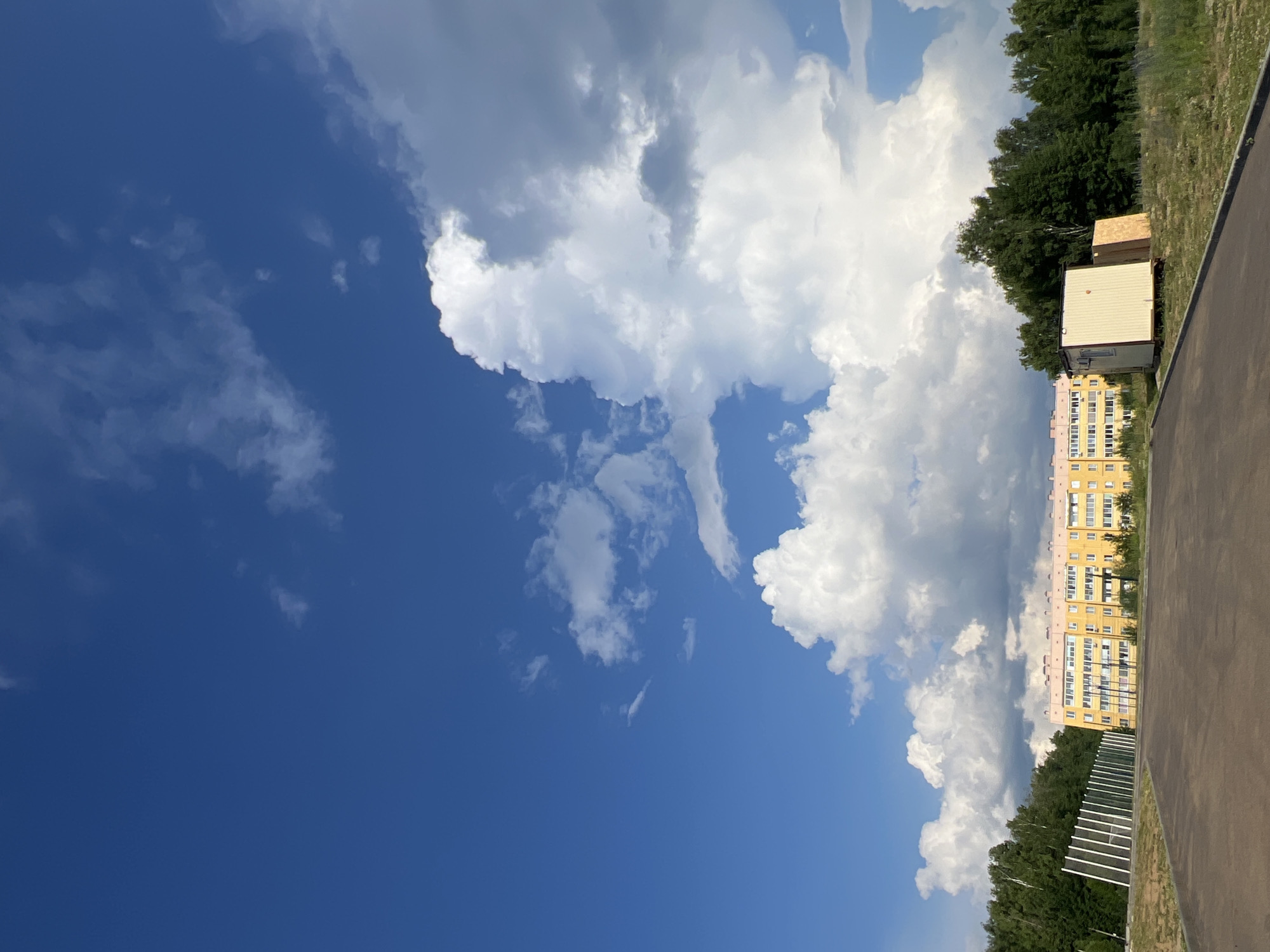 The image depicts a bright, overcast day with a vast expanse of blue sky dotted with large, fluffy white clouds. In the foreground, a paved road curves gently. To the left of the road is a low, green grassy area bordered by a white fence. Further back, a long, yellow, multi-story building stands out against the greenery of a dense forest. To the right of the building, a small, portable office structure is visible, along with some lumber. The overall scene suggests a developing urban or suburban area on the outskirts of Noviy, Russia, with construction or renovation activities ongoing. There are no people or animals visible in the image.