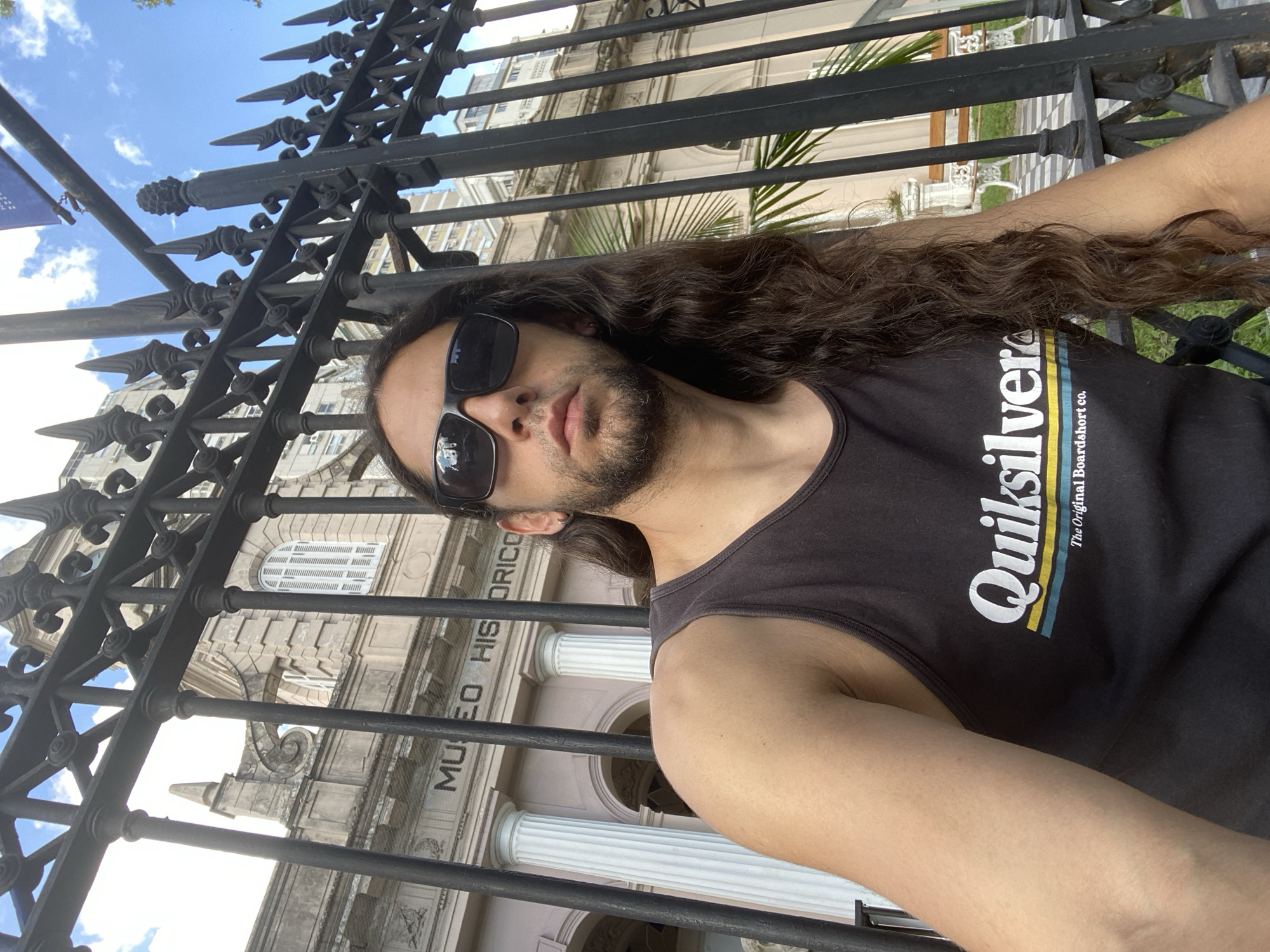 A selfie shot shows a man with long brown hair and sunglasses in front of a black wrought iron fence. The man is wearing a black sleeveless shirt with "Quiksilver" printed on it. Behind the fence, a pale pink building with white columns is visible. The sign "Museo Historico" is visible on the building. The scene is in Buenos Aires, Argentina. The sky is partly cloudy.