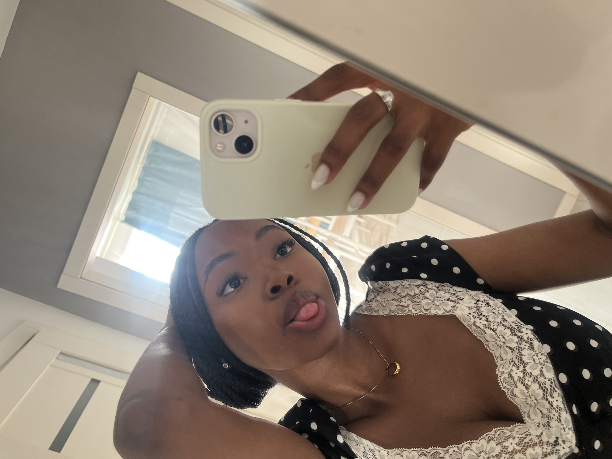 A young woman is taking a selfie in a mirror. She has dark braided hair and is wearing a black and white polka dot dress with white lace trim. She is sticking her tongue out and her left hand is resting on her head. Her right hand is holding a light green phone. She is wearing a gold pendant necklace and a silver ring on her left ring finger. The background appears to be a home interior with a doorway and a mirror reflecting part of a room. There is no discernible connection to Kazan, Russia.