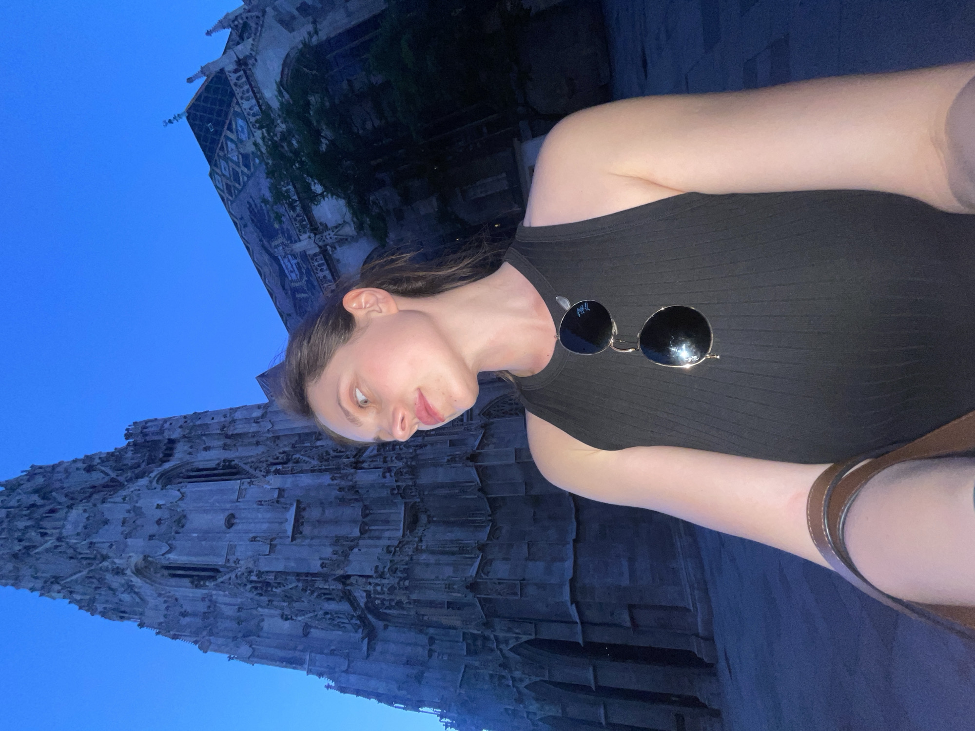 A vertically oriented, low-angle selfie shows a person standing near St. Stephen's Cathedral in Vienna, Austria. The person, a young woman with fair skin, brown hair pulled back, and wearing a black tank top with sunglasses clipped to the front, is facing the camera. The top of the cathedral, a large Gothic structure, dominates the background, rising into a blue sky. The building's intricate stonework is visible. The person holds a brown strap of some sort over their shoulder. The lighting is soft, suggesting it's dusk or dawn.