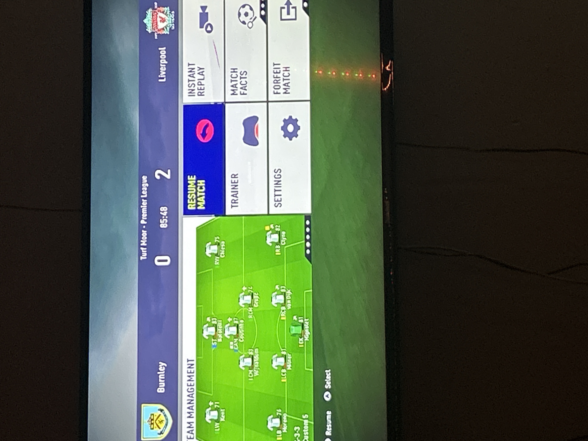 The image is a photograph of a television screen displaying a menu from the FIFA video game. The game shows a match between Burnley and Liverpool in the Premier League, with Liverpool leading 2-0 in the 85th minute. The menu includes options for resuming the match, viewing an instant replay, accessing the trainer, viewing match facts, adjusting settings, and forfeiting the match. Also visible on the screen is a team management display, showing the player formations for Liverpool. There are no people visible in the image, only the television screen and a dark background. There is no discernible connection to the city of Kalitinovo, Bulgaria.