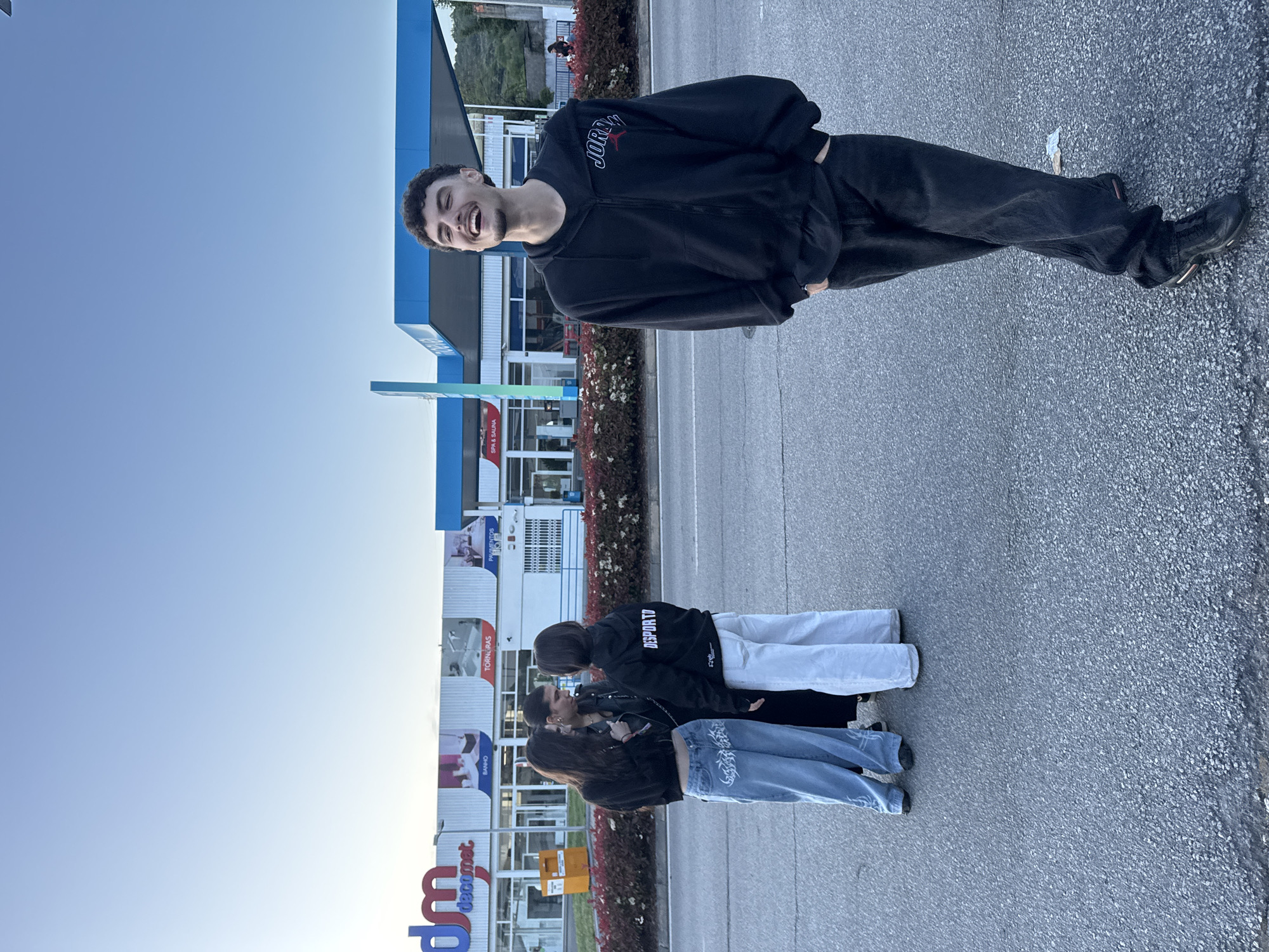 The image captures a group of young people in front of a building. The individuals are assembled in a row. In the background is a building with large signs, including one that reads "bm" and "decomet." The building appears to be a commercial establishment. The setting seems to be an urban area, possibly a parking lot or the street in front of the building. The sky is clear and the lighting suggests it is daytime. The location is likely Bragança, Portugal, based on the signage in the background.
