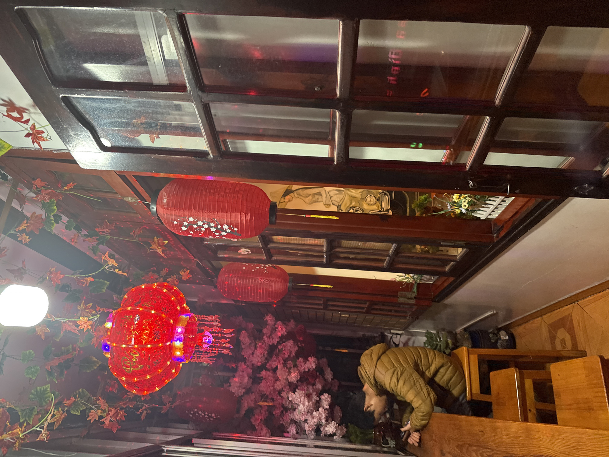 This image depicts the interior of a cafe or restaurant in Da Lat, Vietnam, characterized by a vibrant and warm atmosphere. A single individual, dressed in a mustard-yellow puffer jacket, is seated on a wooden stool at a long wooden counter on the left side, focused on a mobile device held in their hands. The ceiling is extensively decorated with artificial green and autumnal red/orange foliage. Suspended from the ceiling are a bright white overhead light and a large, ornate red lantern featuring gold script, multiple dangling tassels, and embedded multicolored lights. Several smaller, cylindrical red paper lanterns adorned with white cherry blossom patterns are also visible, hanging along the right side of the frame.
The right side of the room features dark wooden window frames with multiple panes, reflecting interior lights and indistinct shapes. Through the glass, elements of the outdoor or adjacent space are subtly visible, including a section of a mural depicting a person in a traditional Vietnamese conical hat (nón lá) next to a vintage vehicle, and a small white picket fence with artificial yellow flowers. A large cluster of artificial pink cherry blossoms adds a prominent decorative feature in the background. The floor is tiled with a light brown geometric pattern, and various potted green plants are placed at floor level along the wall.