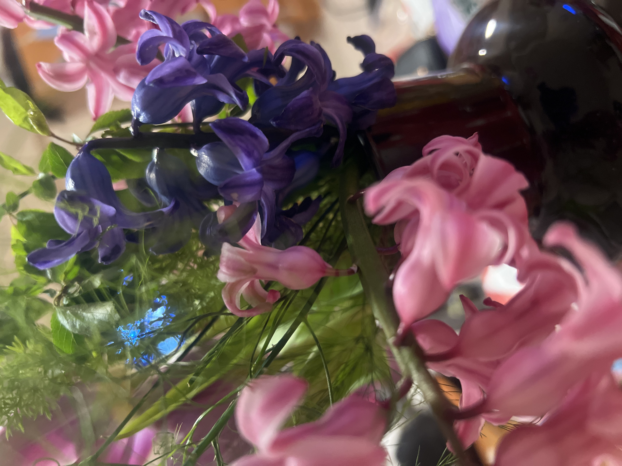 The image shows a close-up of a bouquet of pink and purple hyacinths. There are also some green leaves and stems visible. The background is blurry, but it appears to be a dark, possibly reflective surface, suggesting a vase or container. No location can be determined from this image. No people are visible.